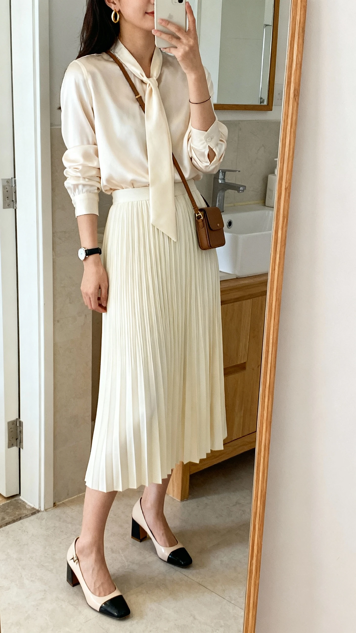 iPhone side-angle mirror selfie of a woman wearing a pleated midi skirt in cream with a silk tie-neck blouse, block-heel slingbacks, minimal gold hoops, a thin watch, and a compact crossbody, face not visible, bathroom mirror with soft indoor lighting, iPhone photo quality.