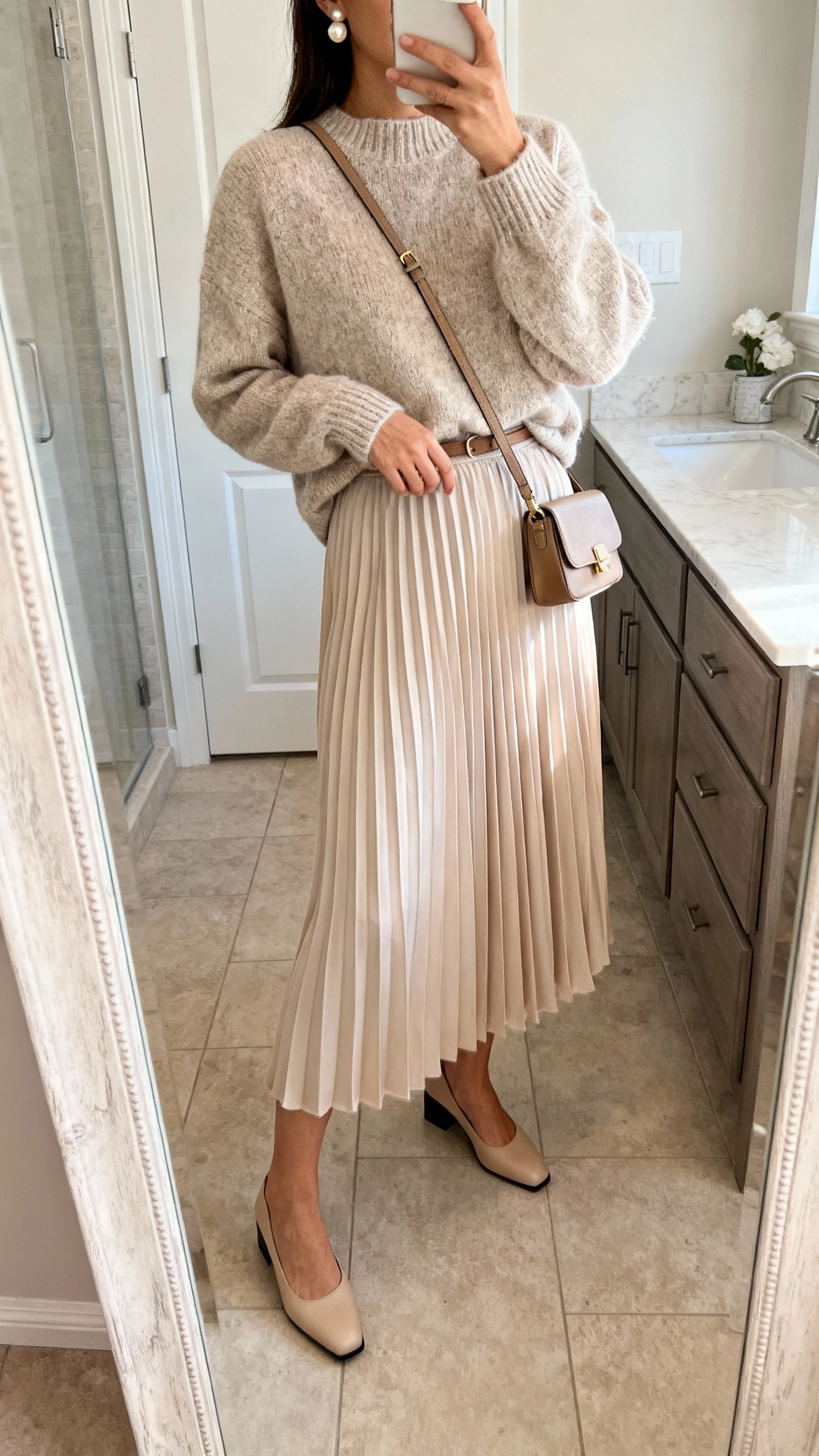iPhone side-angle mirror selfie of a woman wearing a soft knit sweater tucked into a flowy pleated skirt, low block heels, small crossbody bag, and pearl studs, face not visible, bathroom full-length mirror, natural indoor lighting, iPhone photo quality.