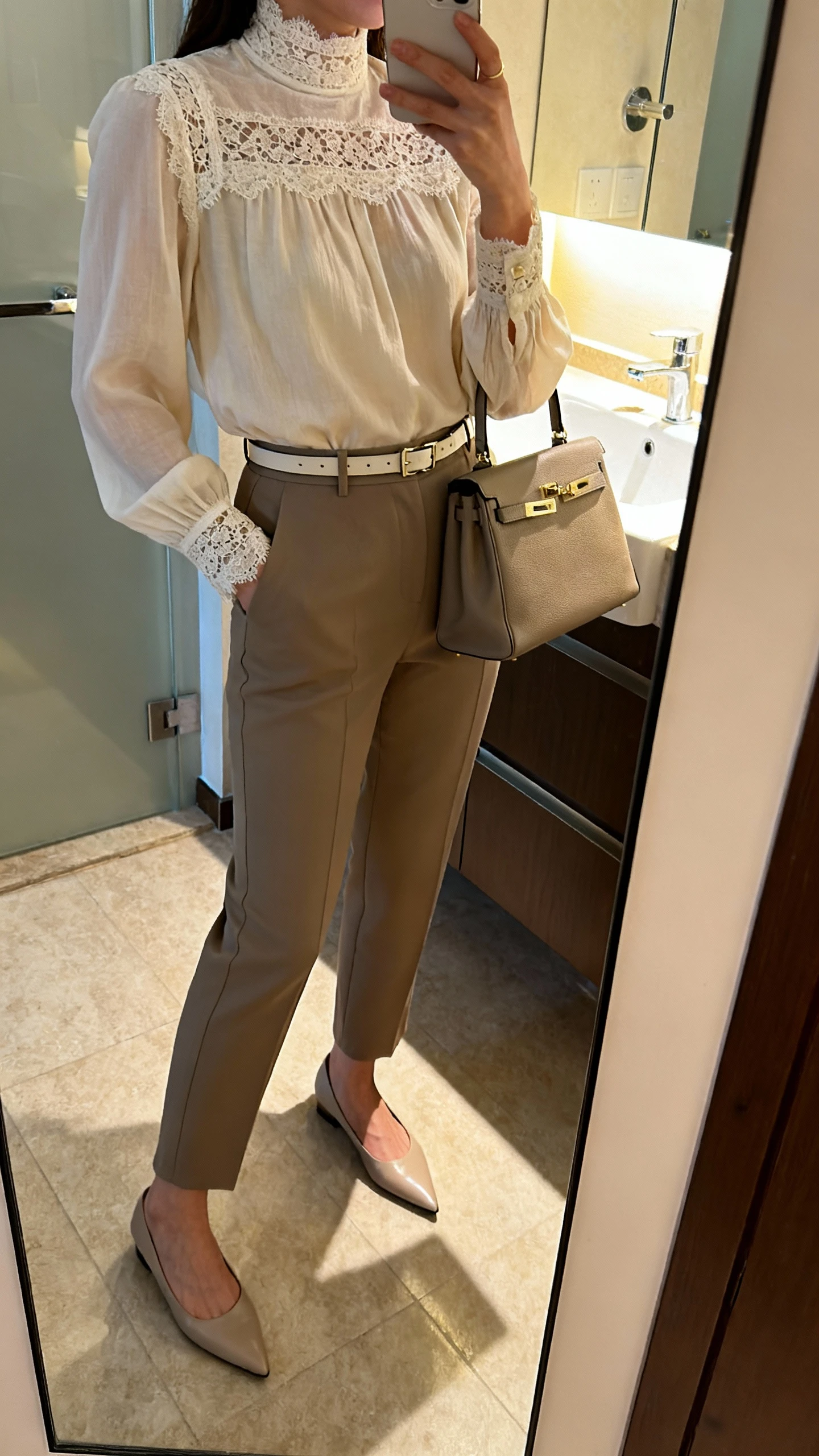 iPhone side-angle mirror selfie of a woman wearing a lace-trimmed high-neck blouse with tailored trousers, slim belt, structured tote, and pointed-toe flats, face not visible, bathroom mirror with indoor lighting, iPhone photo quality.
