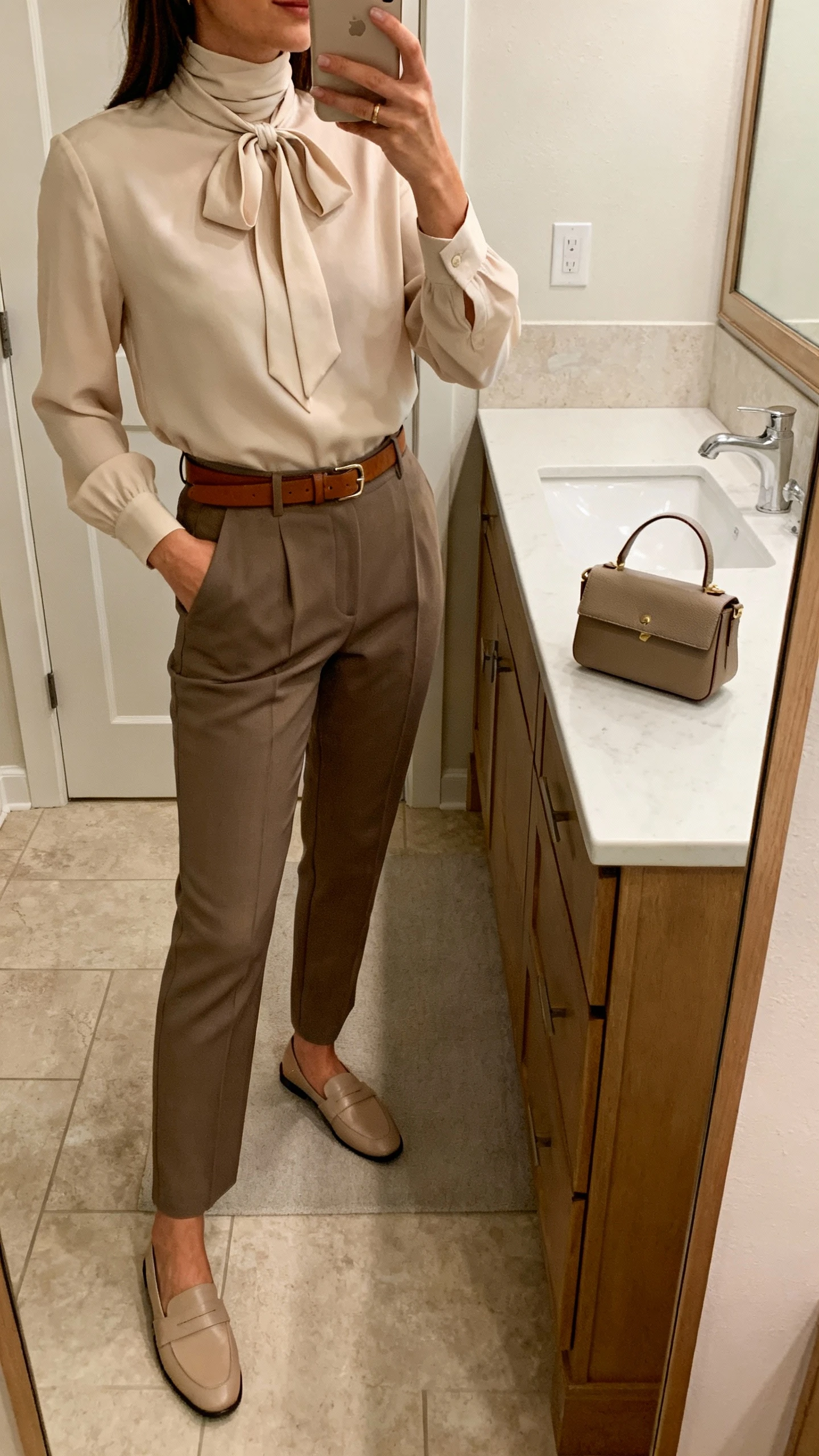 iPhone side-angle mirror selfie of a woman wearing tailored trousers with a bow-neck high-neck blouse, slim leather belt, minimalist loafers, and a small structured tote resting by the mirror, face not visible, bathroom mirror, soft indoor lighting, iPhone photo quality.