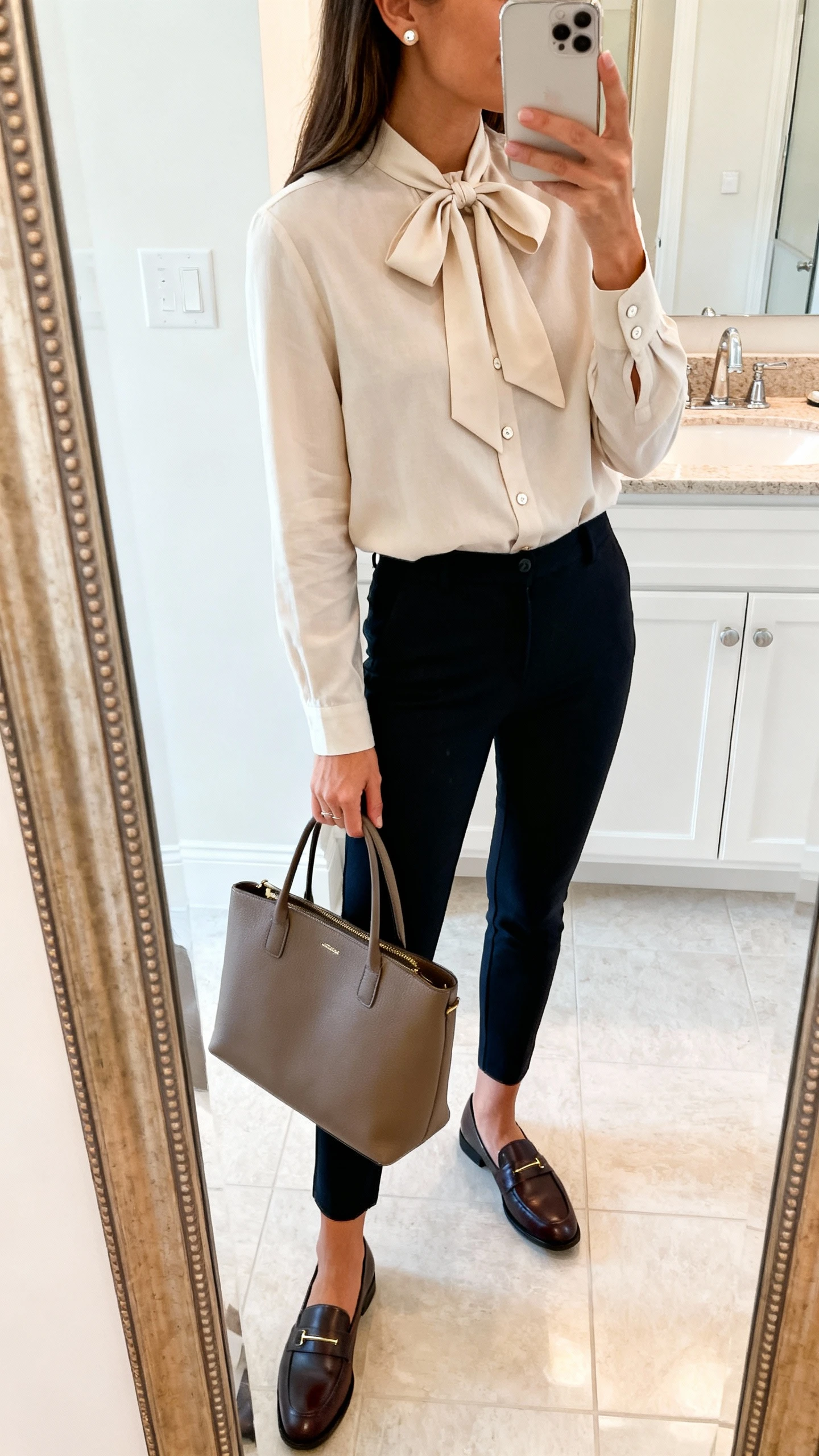 iPhone side-angle mirror selfie of a woman wearing a buttoned-up bow blouse with a tied neck bow, slim ankle pants, structured tote, classic loafers, simple stud earrings, face not visible, bathroom mirror, bright indoor lighting, iPhone photo quality.