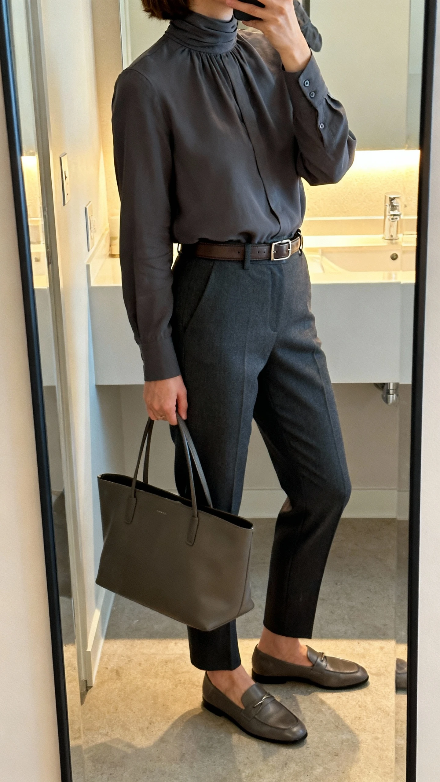 iPhone side-angle mirror selfie of a woman wearing a tailored high-neck blouse tucked into straight-leg trousers in charcoal, slim leather belt, modest loafers, and a structured tote, face not visible, cropped at shoulders, bathroom mirror with soft indoor lighting, iPhone photo quality.