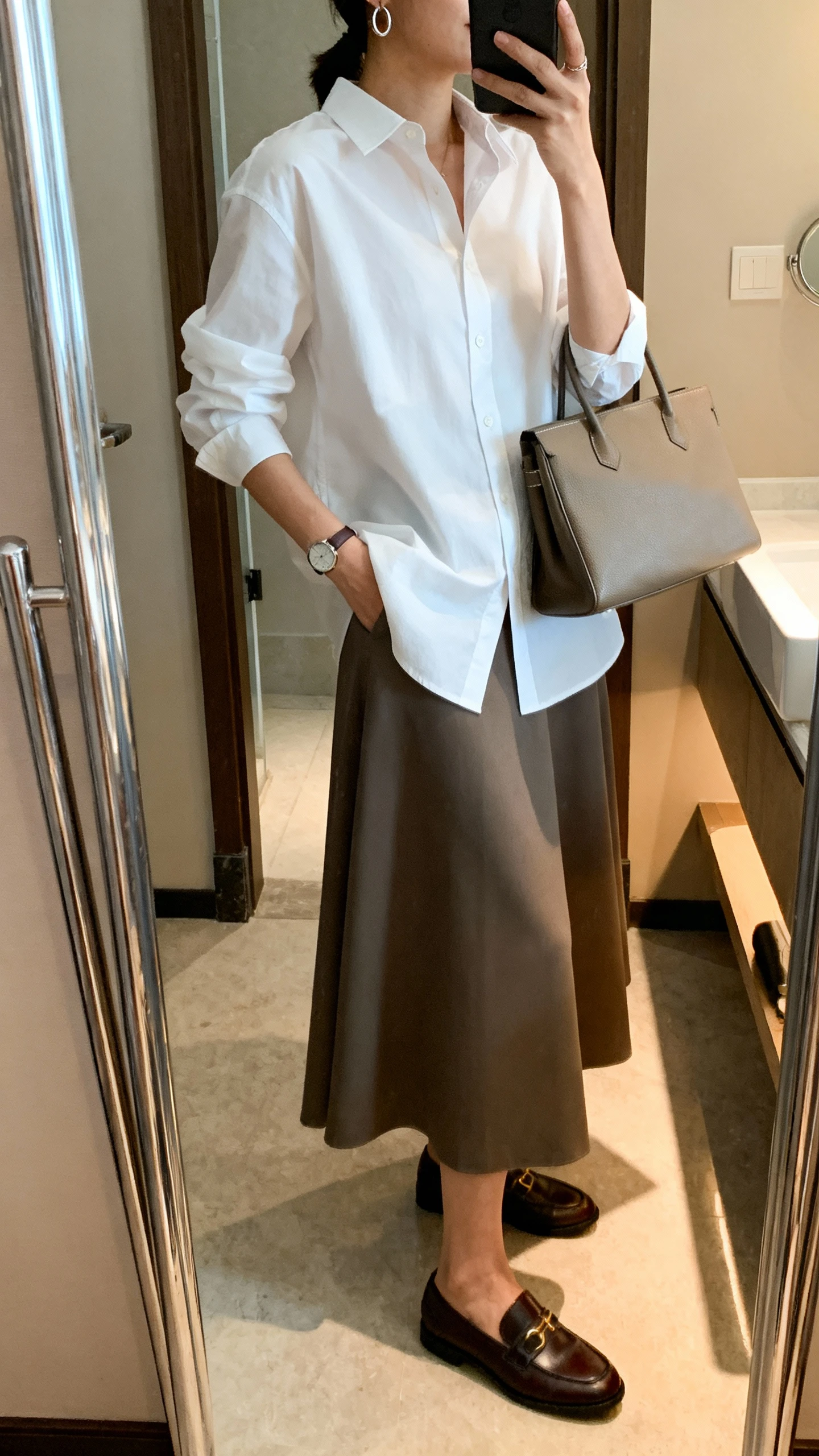 iPhone side-angle mirror selfie of a woman wearing a crisp white button-down shirt with an A-line maxi skirt, classic loafers, a structured tote, a thin watch, and minimal hoop earrings, face not visible, bathroom full-length mirror with indoor lighting, iPhone photo quality.
