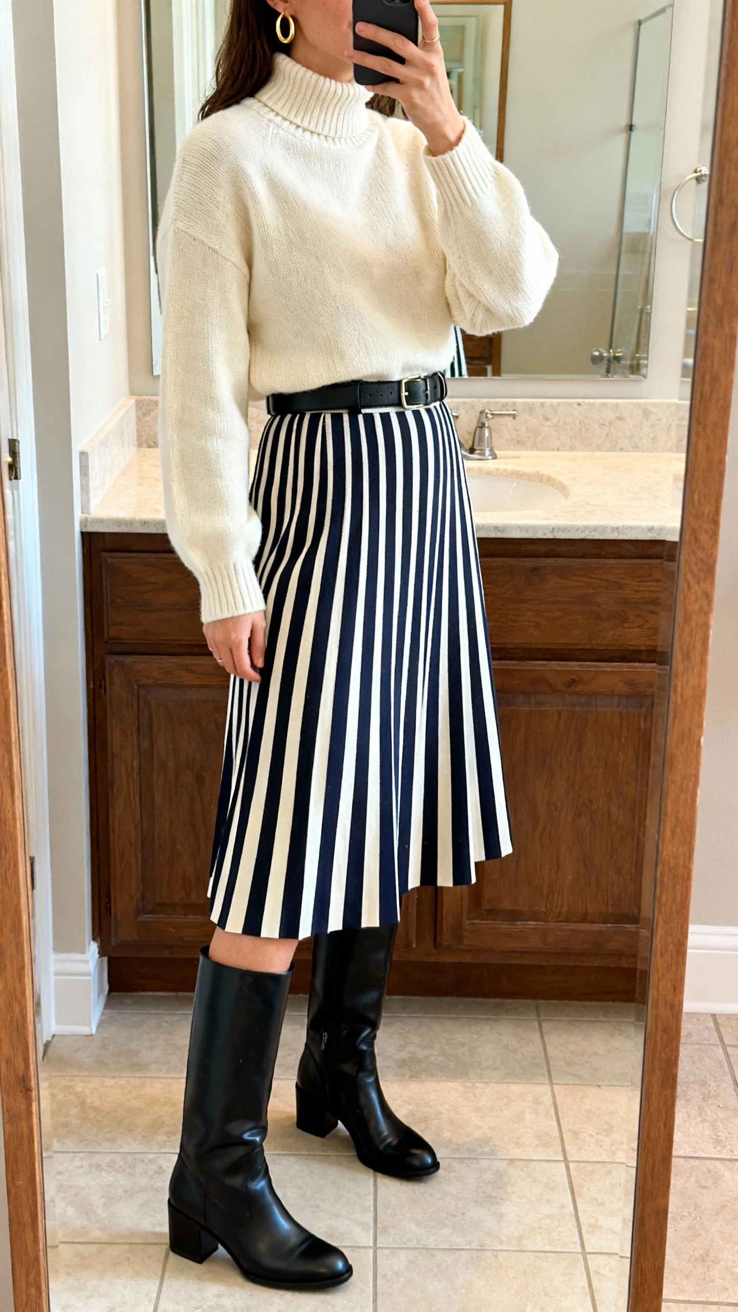 iPhone side-angle mirror selfie of a woman wearing a navy-and-ivory striped A-line skirt (midi), lightweight cream mock-neck sweater, black leather knee-high boots with low block heel, minimal black leather belt, and gold hoop earrings; face not visible, bathroom mirror, morning light, iPhone photo quality.