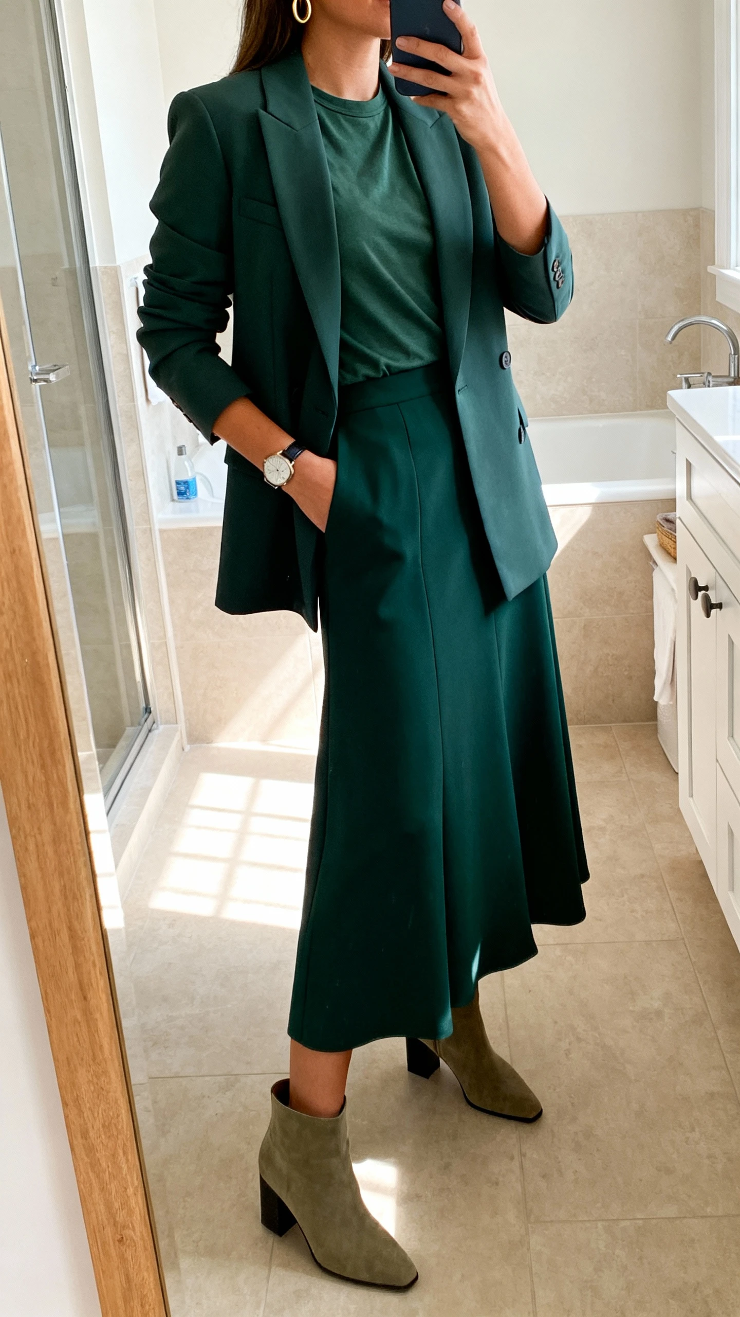 iPhone side-angle mirror selfie of a woman wearing a deep green long A-line skirt, a fitted crewneck tee, a single-breasted blazer in a complementary tone, block-heel ankle boots, minimal gold hoops, and a watch, face not visible, bathroom mirror, indoor morning light, iPhone photo quality.