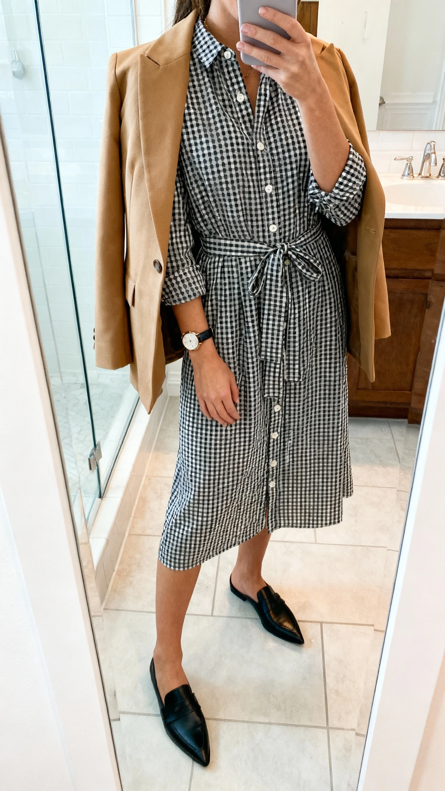 iPhone side-angle mirror selfie of a woman wearing a black-and-white gingham button-front shirtdress with a self-tie belt, structured camel blazer draped over shoulders, pointed black loafers, and a slim leather watch, face not visible, bathroom full-length mirror, bright indoor lighting, iPhone photo quality.