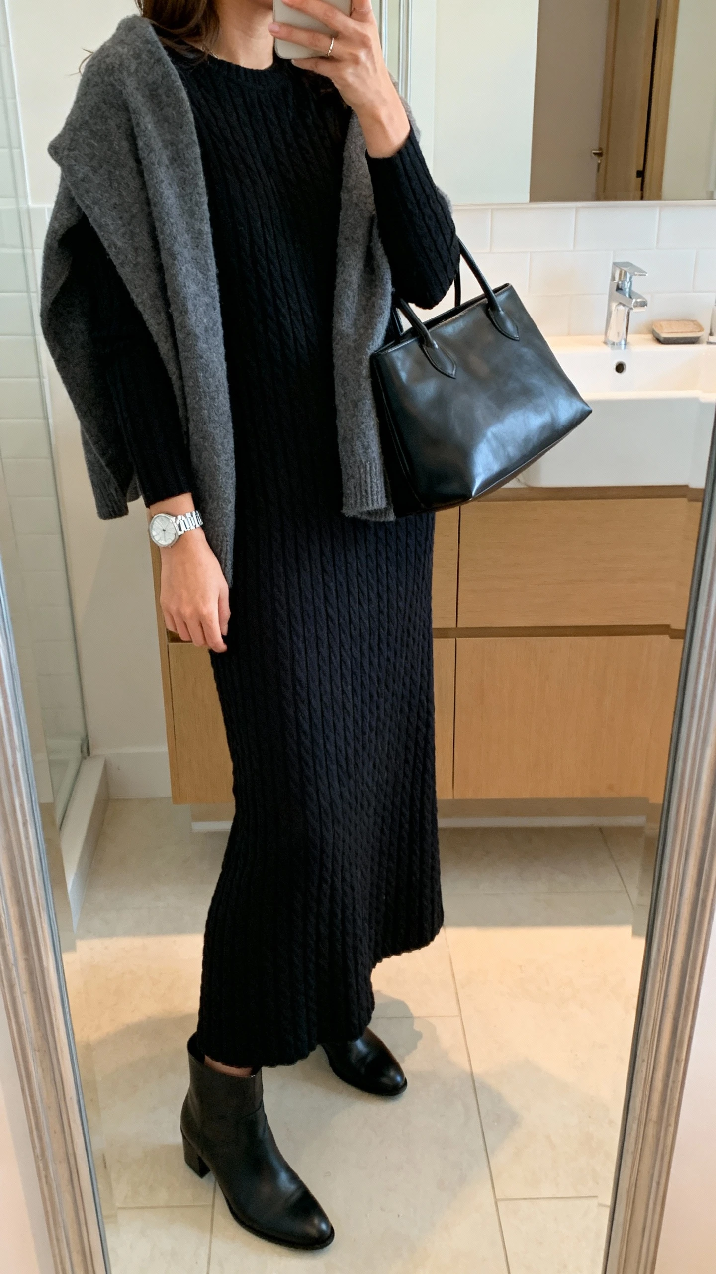 iPhone side-angle mirror selfie of a woman wearing a black ribbed-knit textured maxi dress with long sleeves, a soft charcoal cardigan draped over shoulders, black ankle boots, a black leather tote, and a silver watch, face not visible, bathroom full-length mirror, warm indoor lighting, iPhone photo quality.