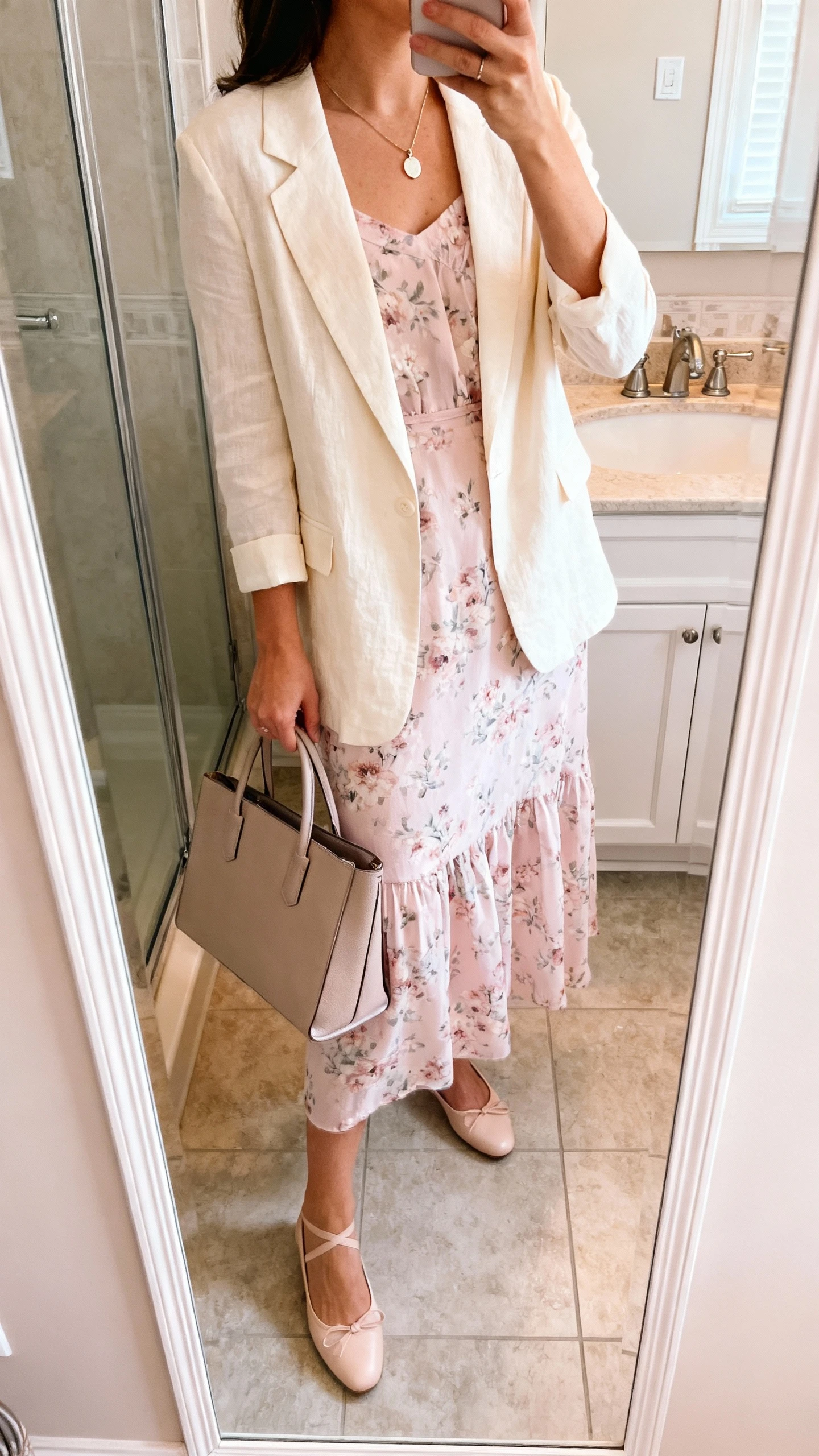 iPhone side-angle mirror selfie of a woman wearing a soft pastel floral dress layered with a lightweight cream blazer, delicate pendant necklace, ballet flats, and a structured tote, face not visible, bathroom full-length mirror, morning indoor lighting, iPhone photo quality.