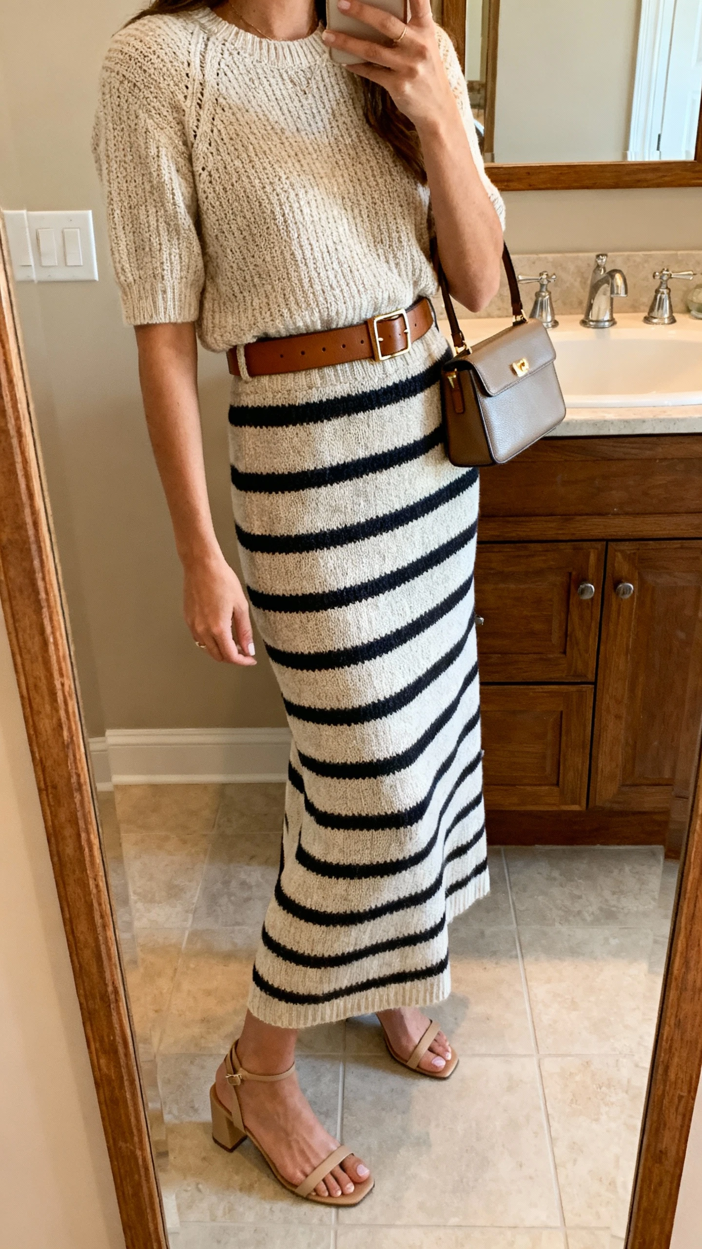 iPhone side-angle mirror selfie of a woman wearing an elevated knit striped maxi skirt with a matching knit top, sleek leather belt, minimalist heeled sandals, structured mini bag, face not visible, bathroom mirror, warm indoor lighting, iPhone photo quality.