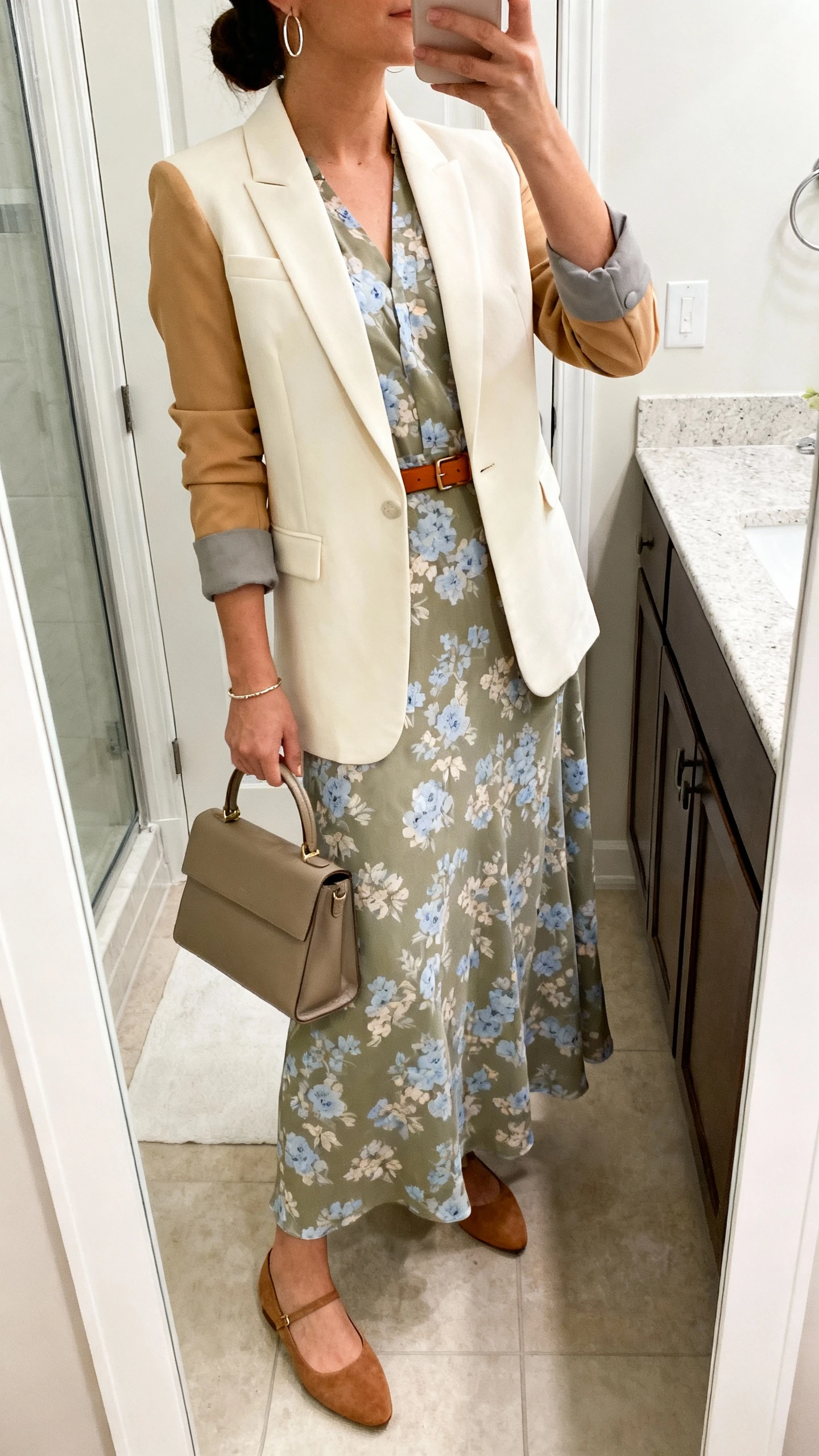 iPhone side-angle mirror selfie of a woman wearing a muted floral maxi dress (sage/soft taupe/powder blue flowers) with a tailored blazer (cream/camel/stone, slim lapel), ballet flats (leather or suede, almond toe), a thin leather belt (tan or chocolate), a structured tote or top-handle bag, and minimal hoop earrings with a simple bracelet, face not visible, bathroom mirror, soft indoor lighting, iPhone photo quality.