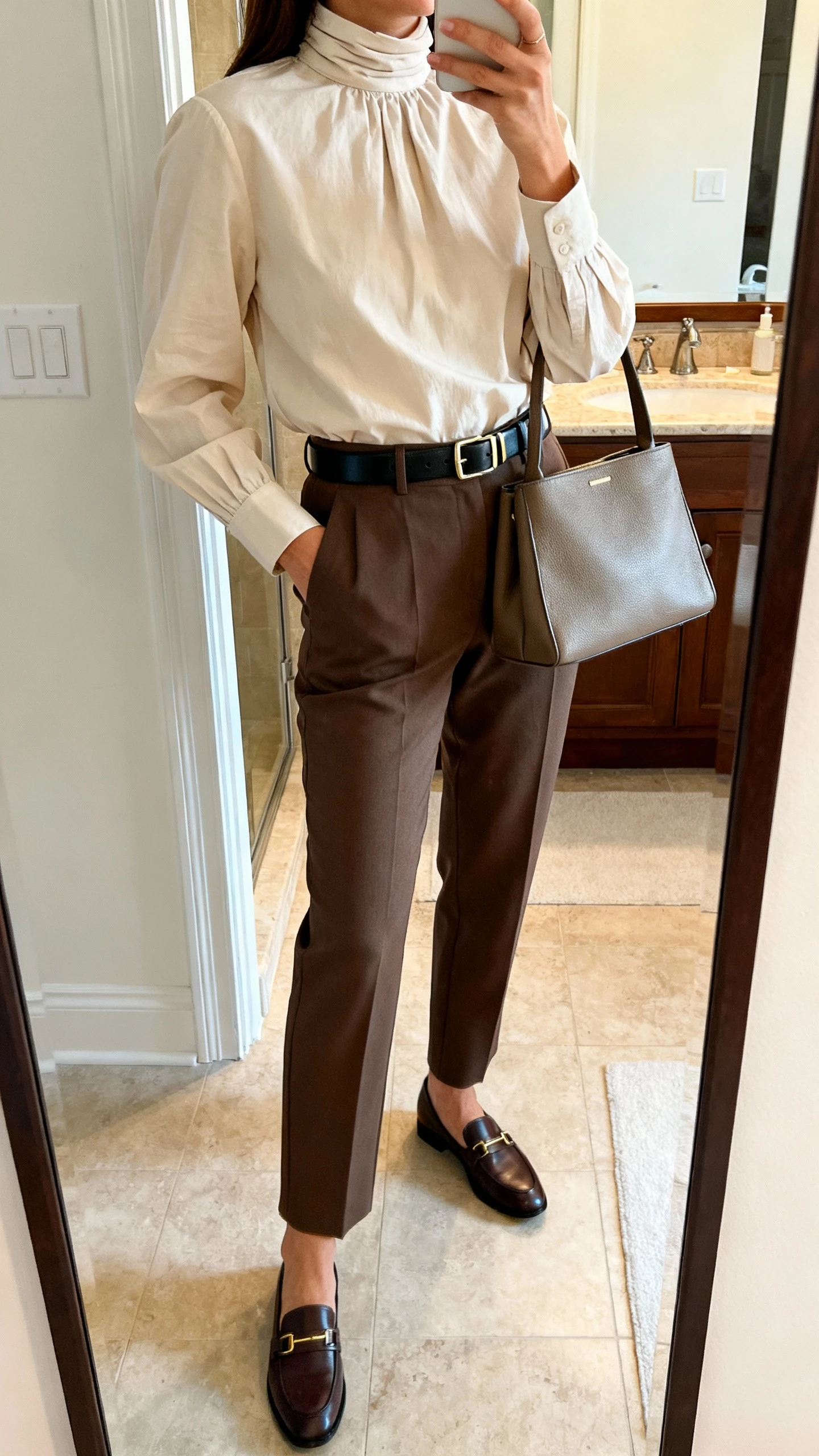 iPhone side-angle mirror selfie of a woman wearing a high-neck cotton blouse with tailored trousers, sleek belt, loafers, and a structured tote, face not visible, bathroom mirror, morning indoor lighting, iPhone photo quality.