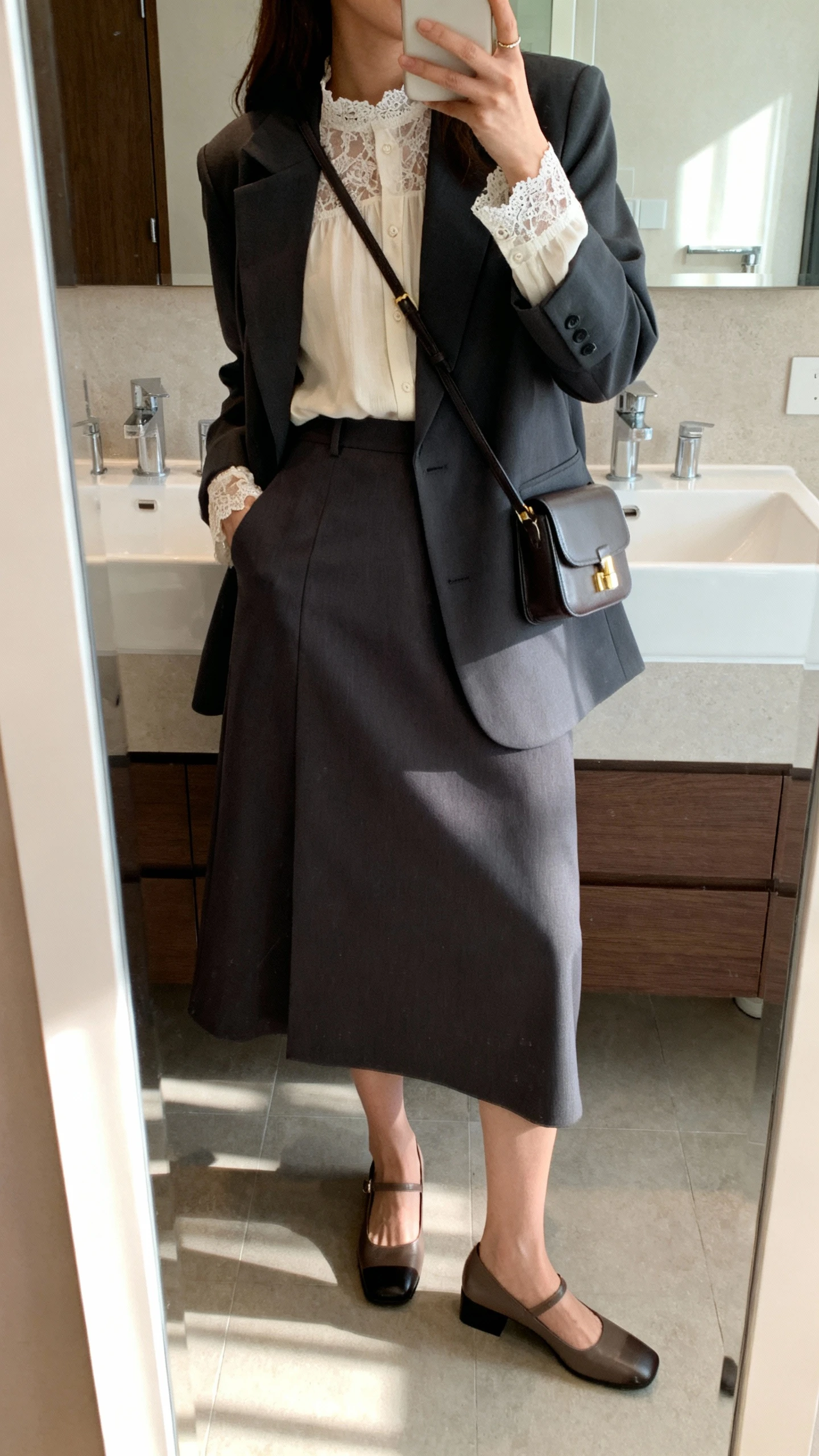 iPhone side-angle mirror selfie of a woman wearing a lace-trim long-sleeve blouse tucked into a midi skirt with a neat structured blazer layered on top, small crossbody bag and low block heels, face not visible, bathroom mirror, indoor natural light, iPhone photo quality.