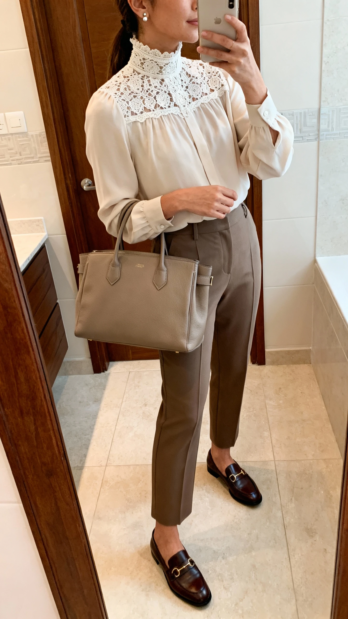 iPhone side-angle mirror selfie of a woman wearing a high-neck lace-collar blouse with tailored trousers, classic loafers, simple stud earrings, and a structured tote; face not visible, cropped at shoulders, bathroom mirror, indoor lighting, iPhone photo quality.