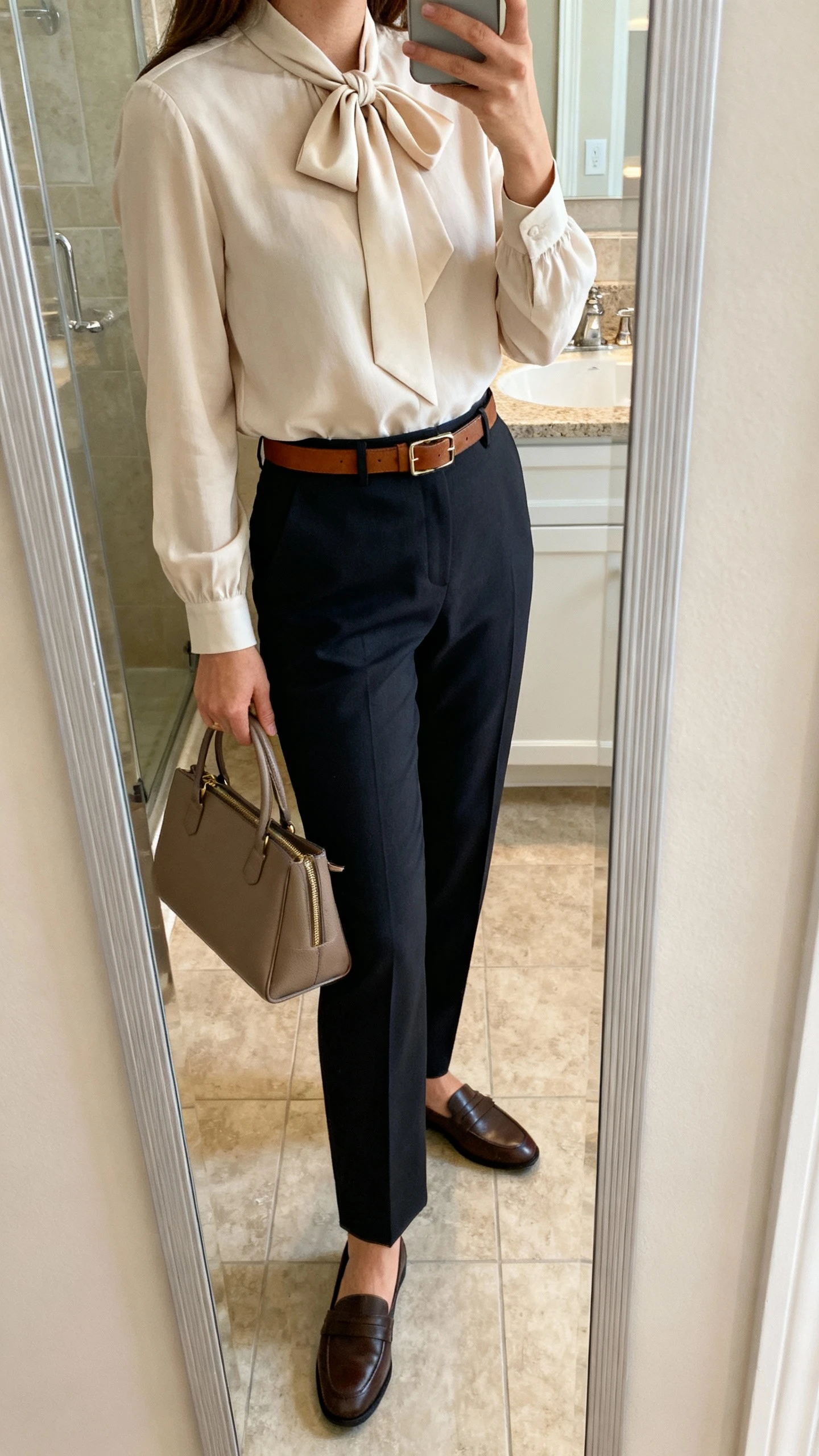 iPhone side-angle mirror selfie of a woman wearing a graceful bow-tie blouse tucked into tailored trousers, with a slim leather belt, structured tote, and loafers, face not visible, bathroom full-length mirror, soft indoor lighting, iPhone photo quality.