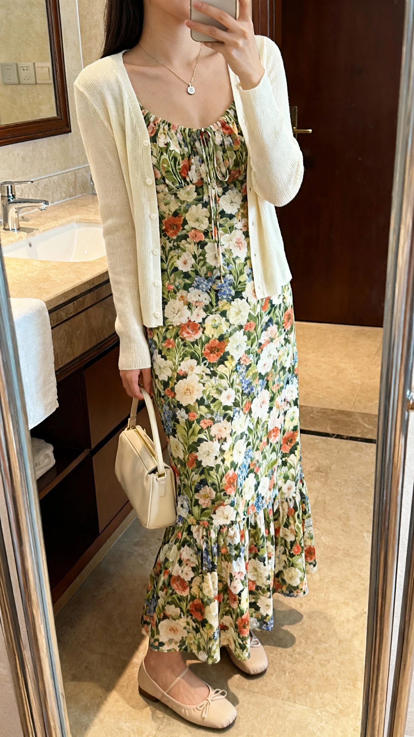 iPhone side-angle mirror selfie of a woman wearing a garden print floral maxi dress with a cream cardigan, delicate pendant necklace, small tote, and flat ballet-style shoes, face not visible, bathroom mirror, indoor warm lighting, iPhone photo quality.
