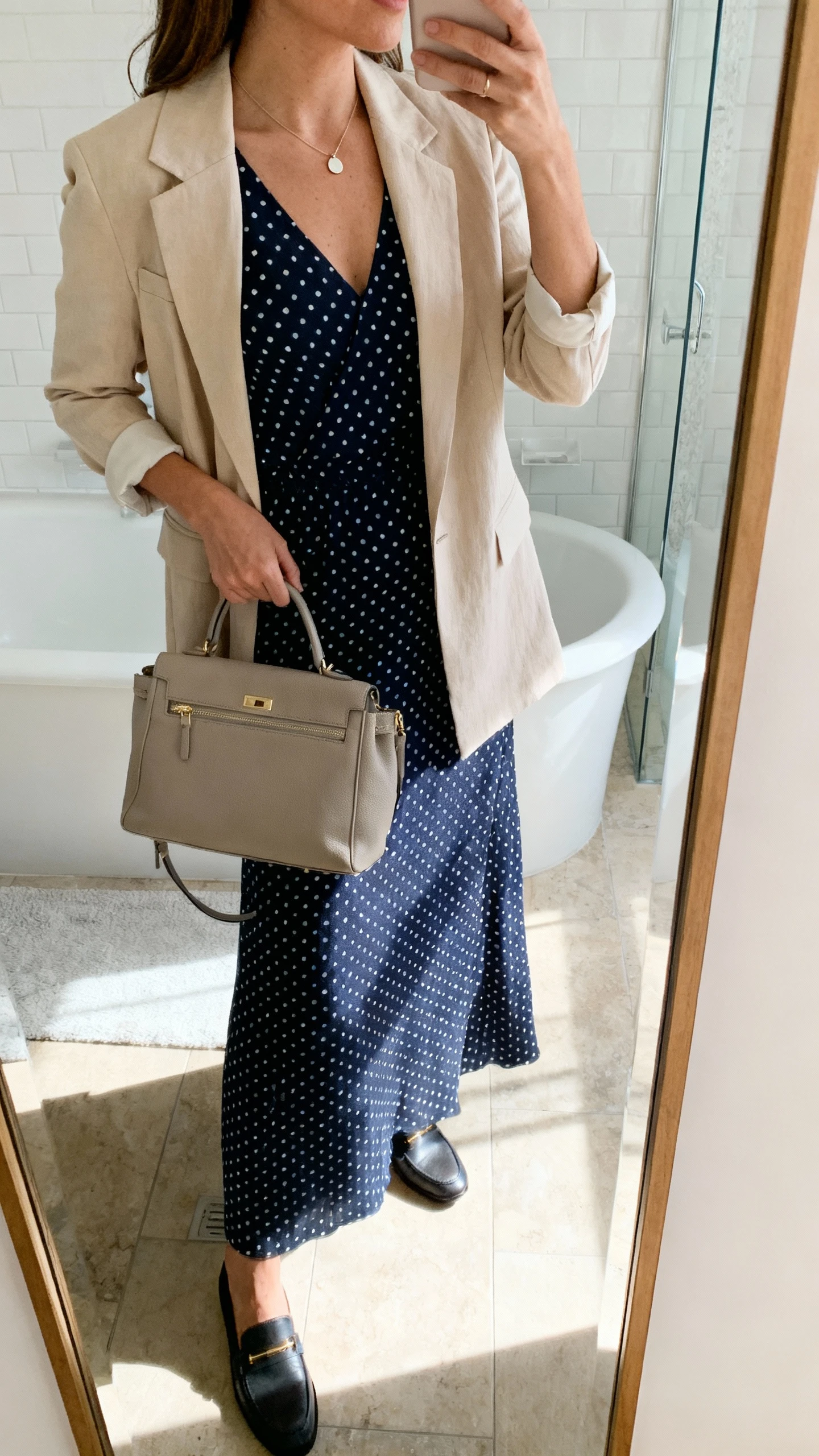 iPhone side-angle mirror selfie of a woman wearing a navy polka dot maxi dress under a soft-shouldered beige blazer, minimal pendant necklace, structured tote, and loafers, face not visible, cropped at shoulders, bathroom mirror, morning light, iPhone photo quality.