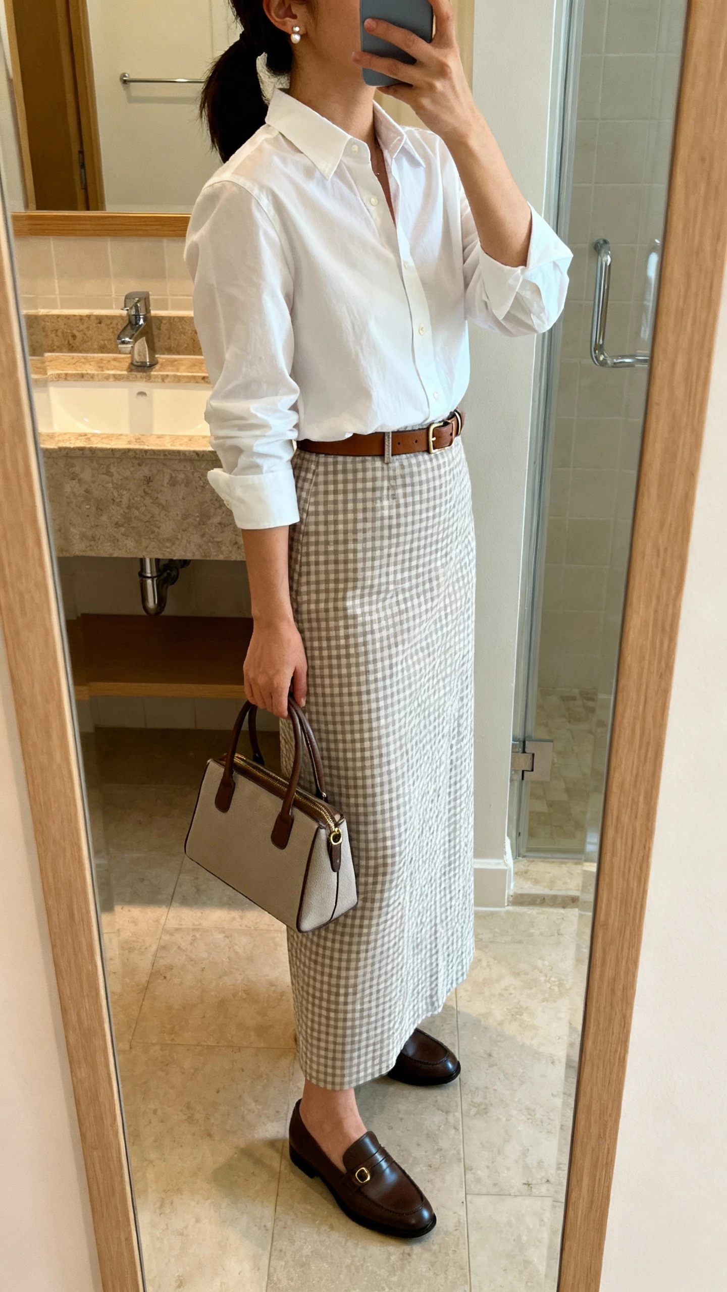 iPhone side-angle mirror selfie of a woman wearing a crisp white button-down tucked into a gingham long skirt, slim leather belt, classic loafers, subtle stud earrings, and carrying a structured top-handle bag; face not visible, bathroom full-length mirror, natural indoor lighting, iPhone photo quality.