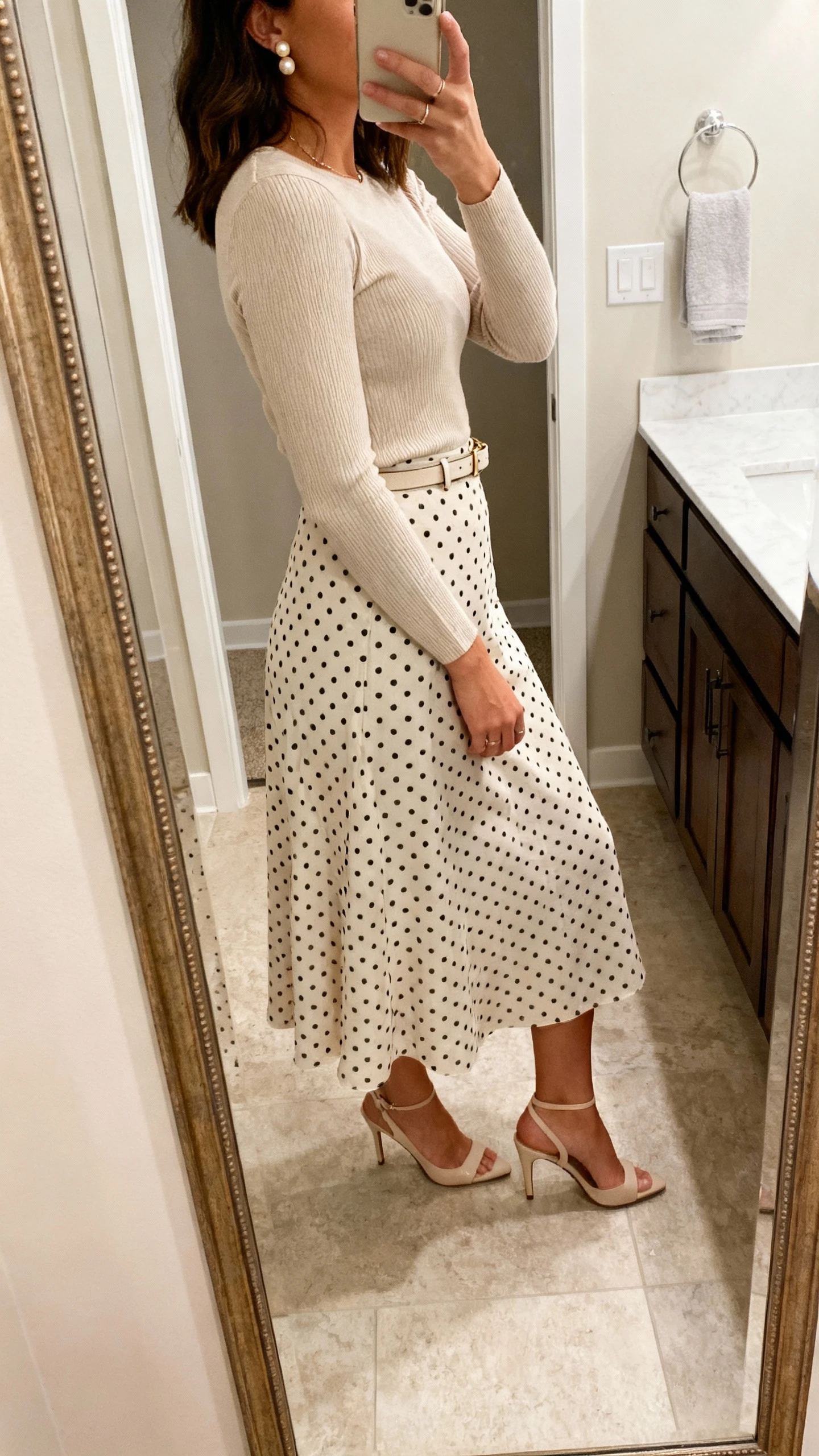 iPhone side-angle mirror selfie of a woman wearing a midi polka-dot skirt, fitted lightweight knit top tucked in, delicate pearl studs, slim belt, and kitten-heel slingbacks, face not visible, bathroom mirror, natural indoor lighting, iPhone photo quality.