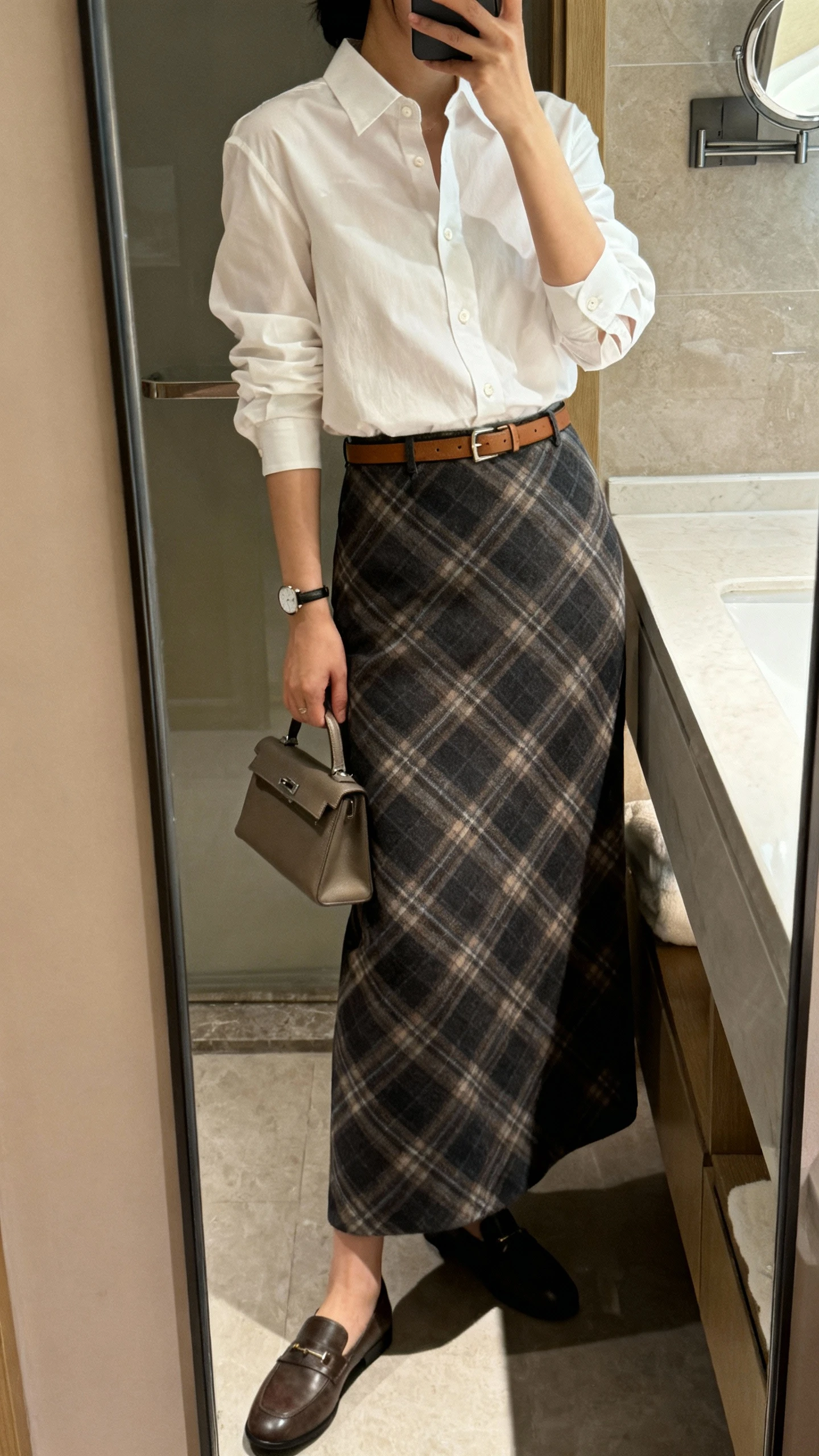 iPhone side-angle mirror selfie of a woman wearing a classic plaid long skirt with a crisp white button-up shirt tucked in, slim leather belt at the waist, loafers, minimal watch, and small structured handbag, face not visible, bathroom full-length mirror, soft indoor lighting, iPhone photo quality.