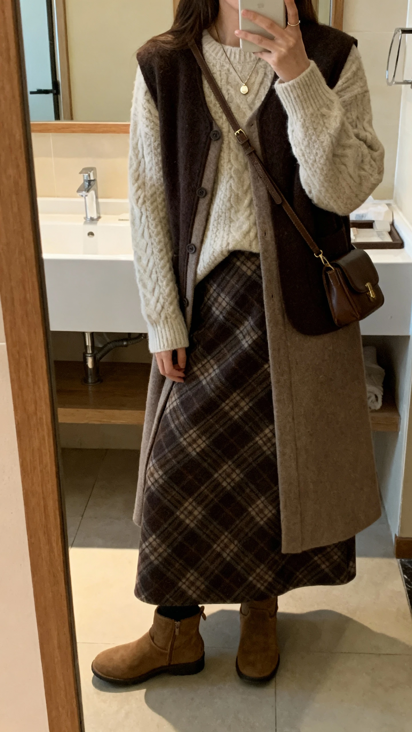iPhone side-angle mirror selfie of a woman wearing a plaid long skirt with a cozy knit sweater and a longline vest layered over, ankle boots, delicate pendant necklace, and a crossbody bag, face not visible, bathroom mirror, indoor warm lighting, iPhone photo quality.