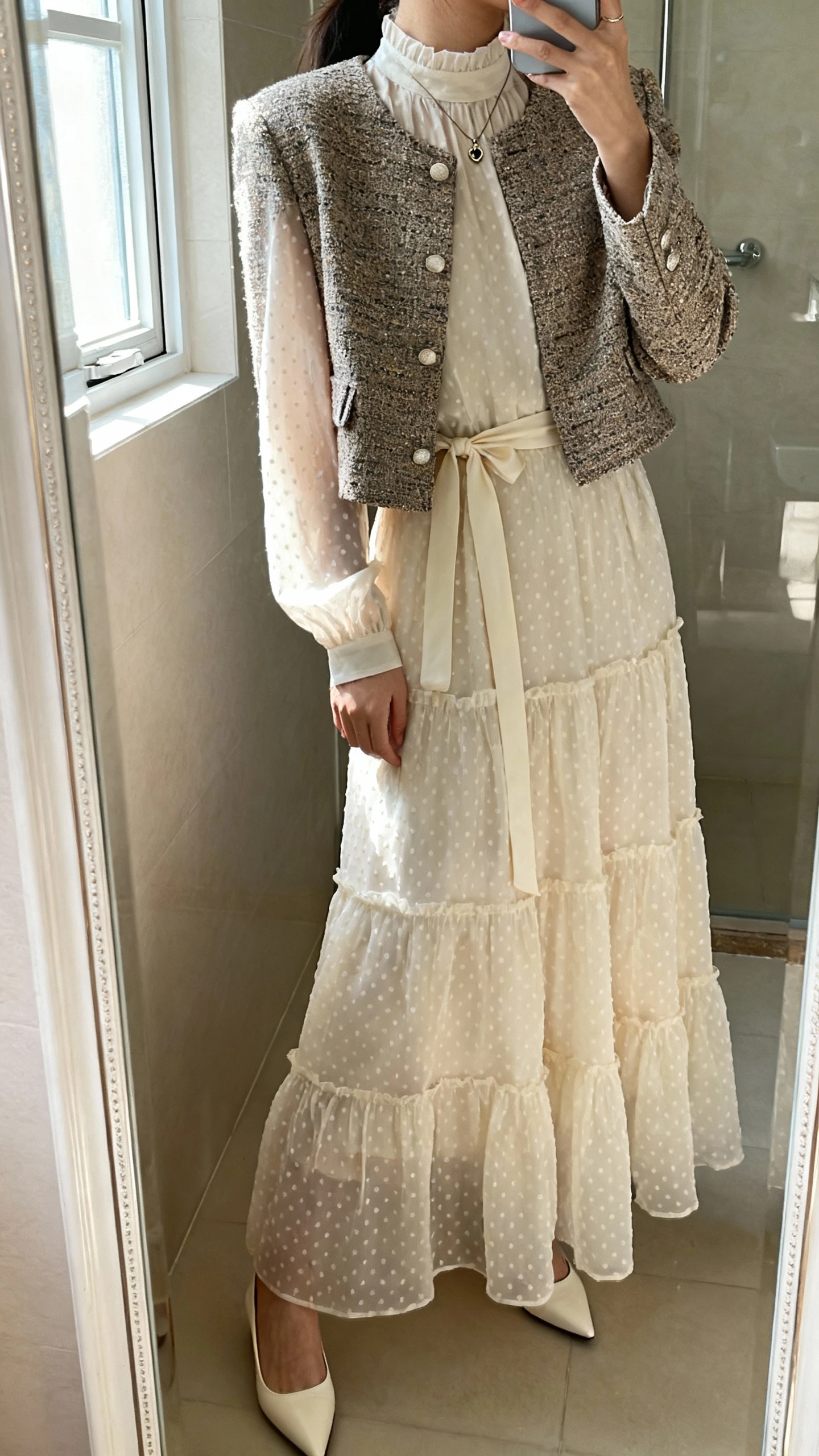 iPhone side-angle mirror selfie of a woman wearing an opaque Swiss dot tiered maxi dress with long sleeves and a high neck, soft waist sash, low pointed-toe flats or kitten heels, a structured blazer or cropped tweed jacket, small pendant necklace worn under the collar; face not visible, bathroom mirror with window light, iPhone photo quality.