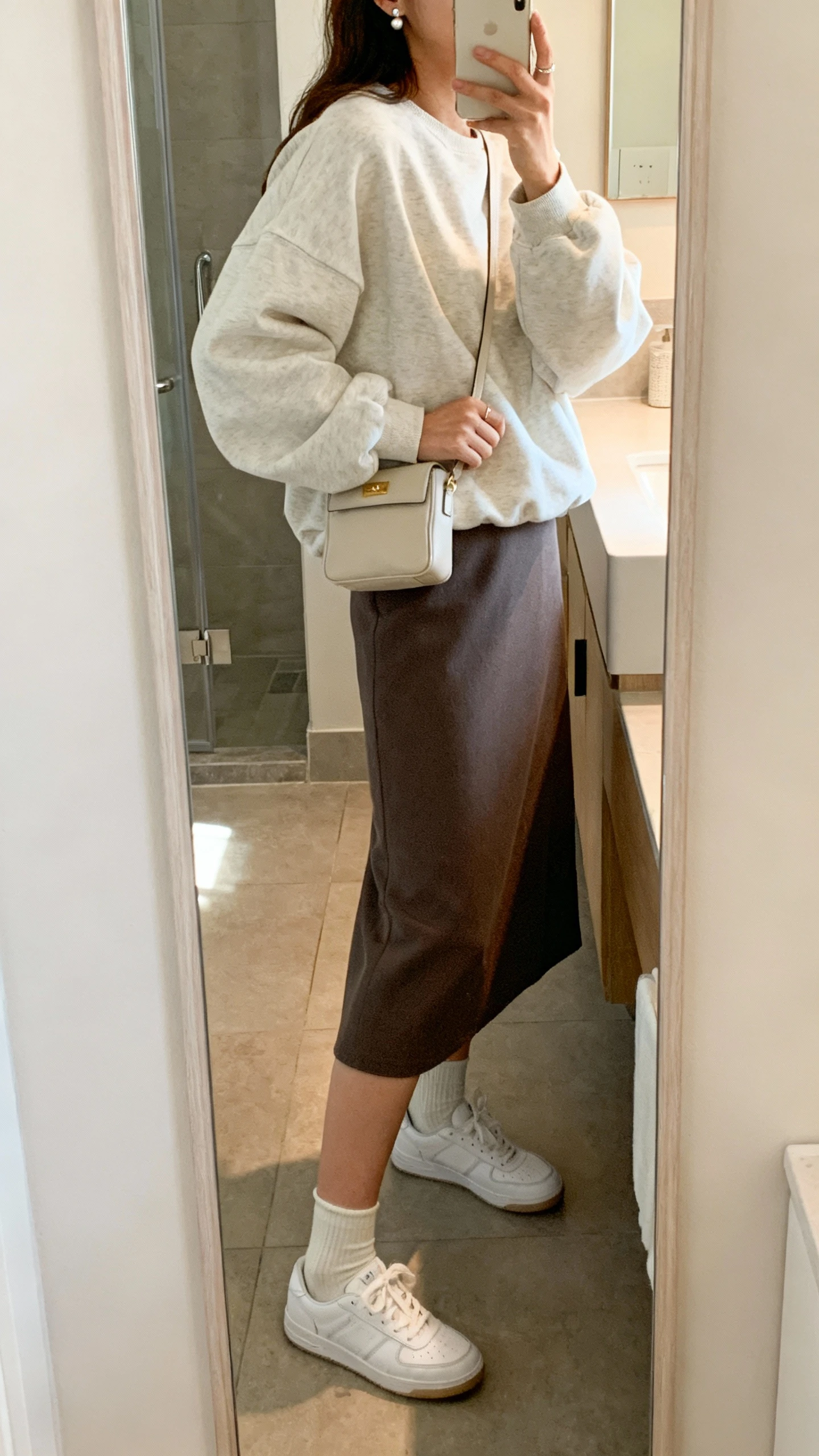 iPhone side-angle mirror selfie of a woman wearing a midi skirt with a slightly oversized sweatshirt, white sneakers, ankle socks, small shoulder bag, dainty studs, face not visible, bathroom full-length mirror, soft morning light, iPhone photo quality.