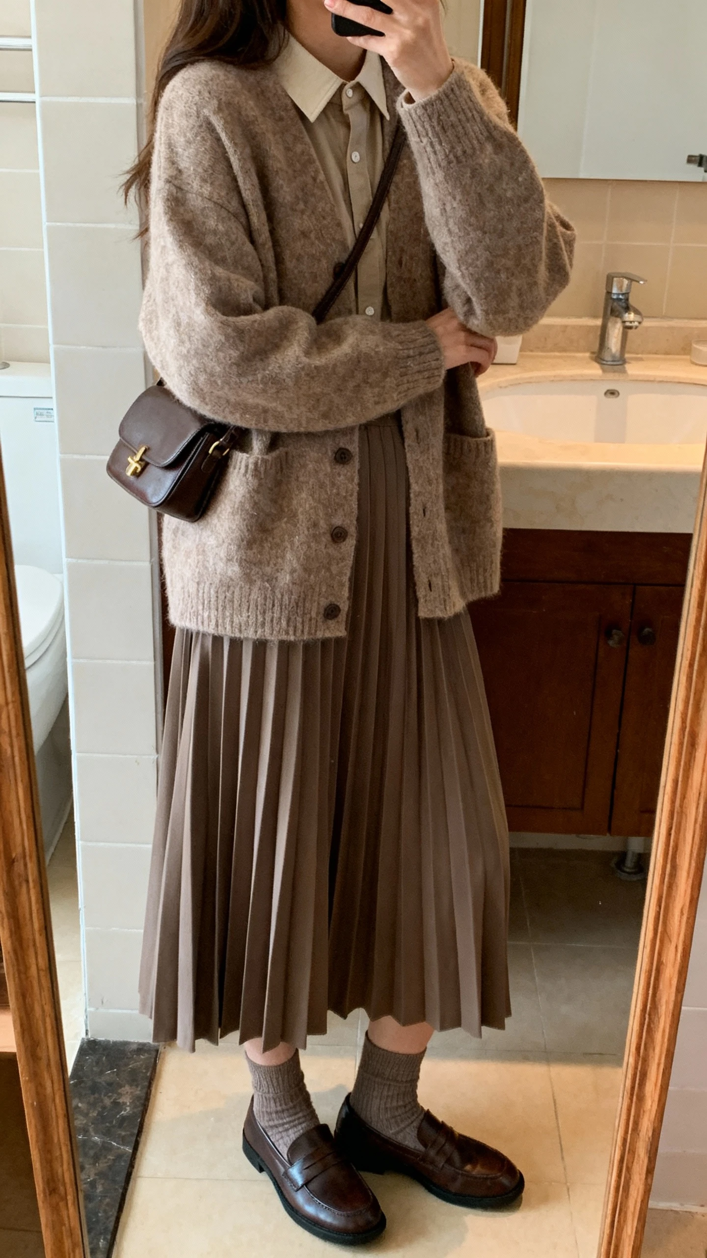 iPhone side-angle mirror selfie of a woman wearing a cozy oversized cardigan layered over a tucked-in top with a flowy pleated midi skirt, ankle socks with loafers, and a small crossbody bag, face not visible, bathroom mirror, warm indoor lighting, iPhone photo quality.
