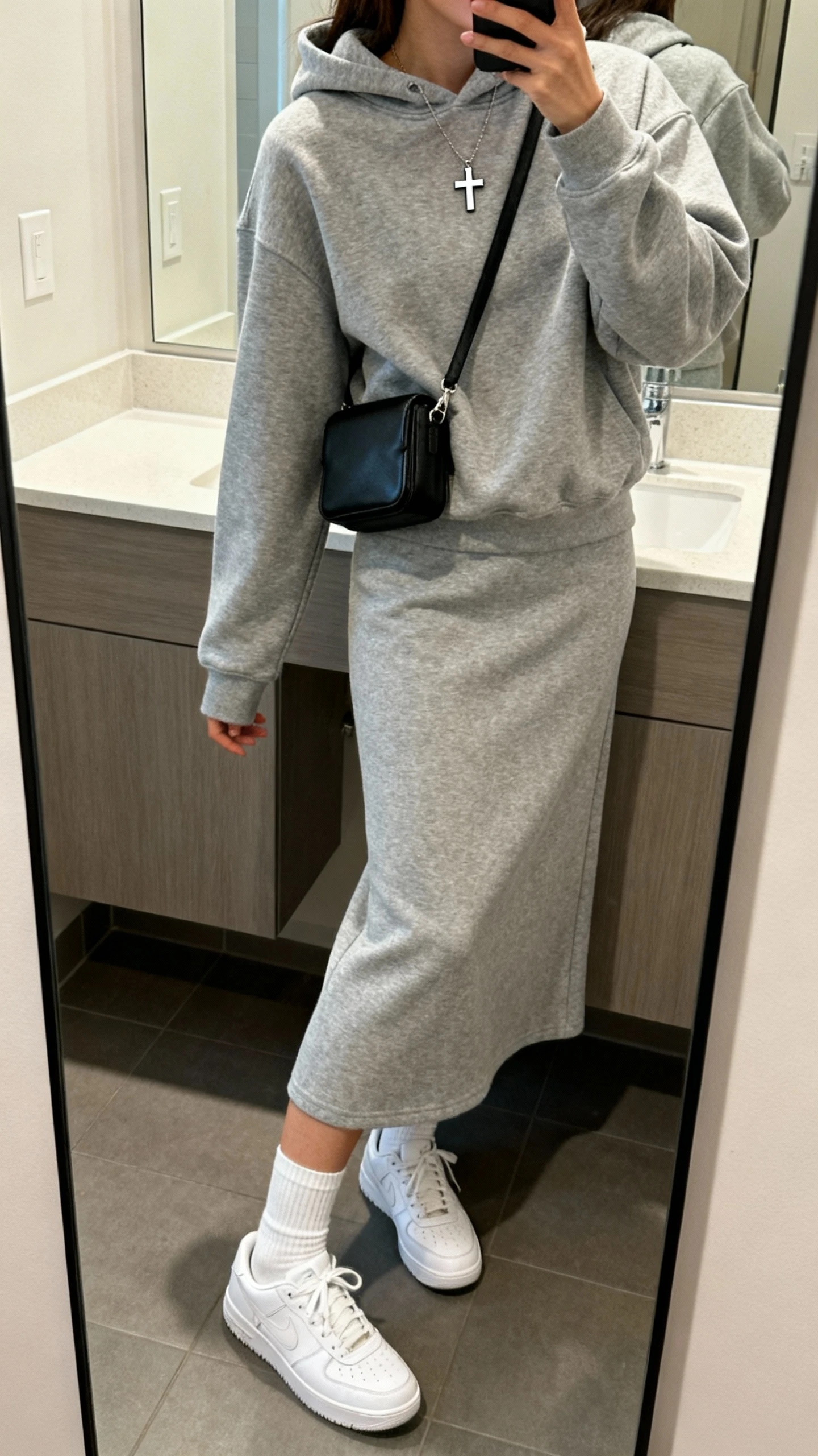 iPhone side-angle mirror selfie of a woman wearing a matching cozy heather gray hoodie and midi skirt set, white crew socks, clean white sneakers, small cross pendant, and a compact black crossbody; face not visible, bathroom mirror, soft indoor lighting, iPhone photo quality.