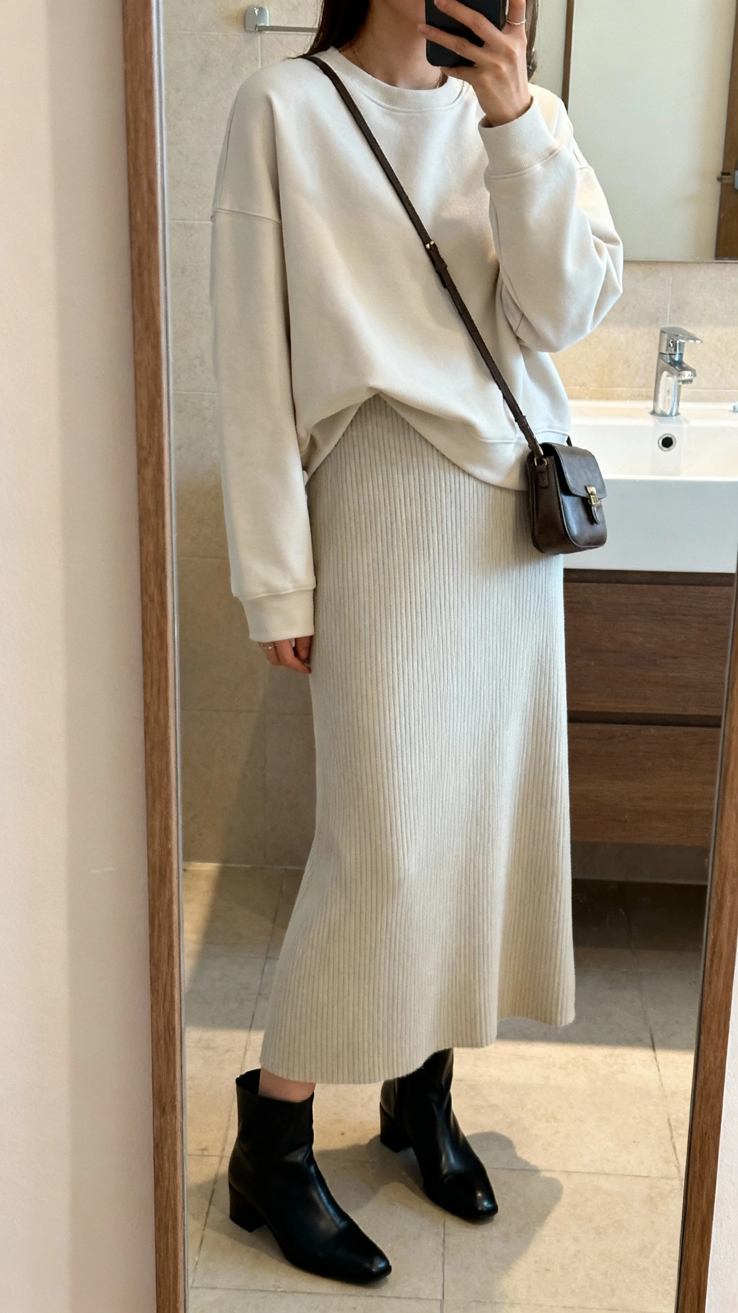 iPhone side-angle mirror selfie of a woman wearing a monochrome minimalist crewneck over a ribbed maxi dress, paired with sleek black ankle boots and a small crossbody bag, face not visible, cropped at shoulders, bathroom mirror, soft indoor lighting, iPhone photo quality