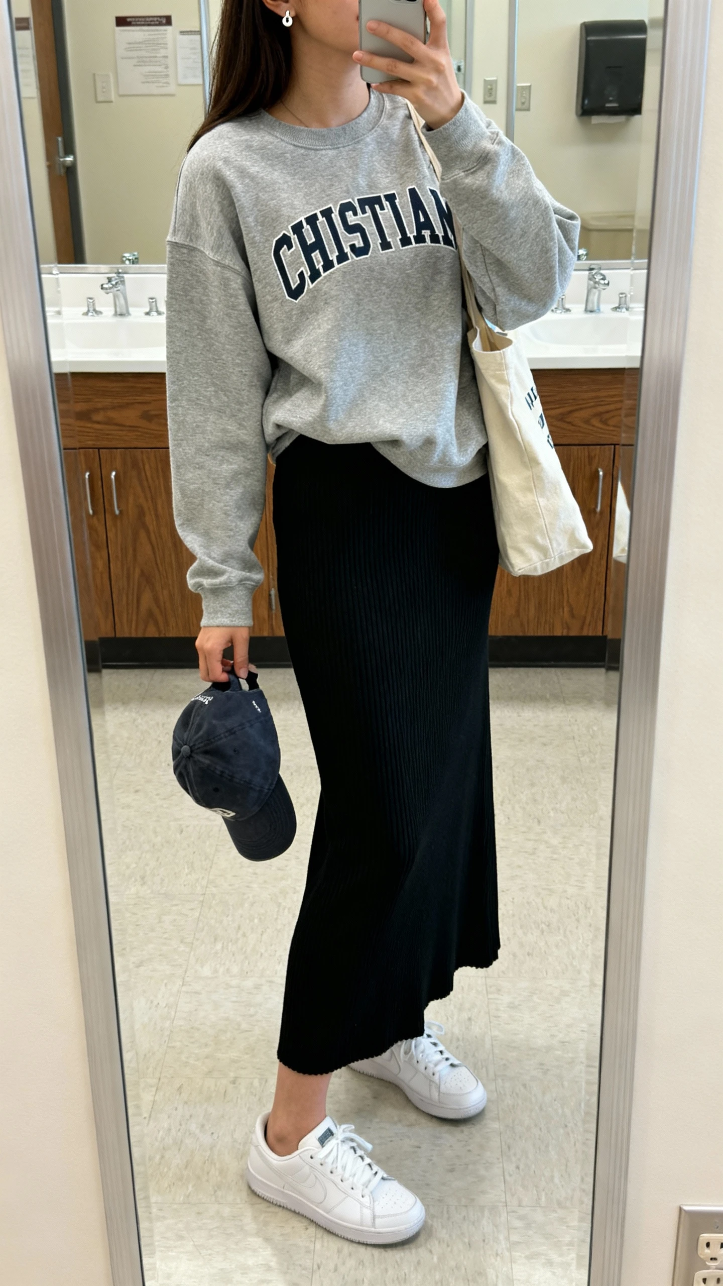 iPhone side-angle mirror selfie of a woman wearing a heather gray Christian crewneck over a ribbed black maxi dress, white sneakers, canvas tote, simple stud earrings, and a baseball cap in hand, face not visible, bathroom mirror, soft indoor campus-style lighting, iPhone photo quality