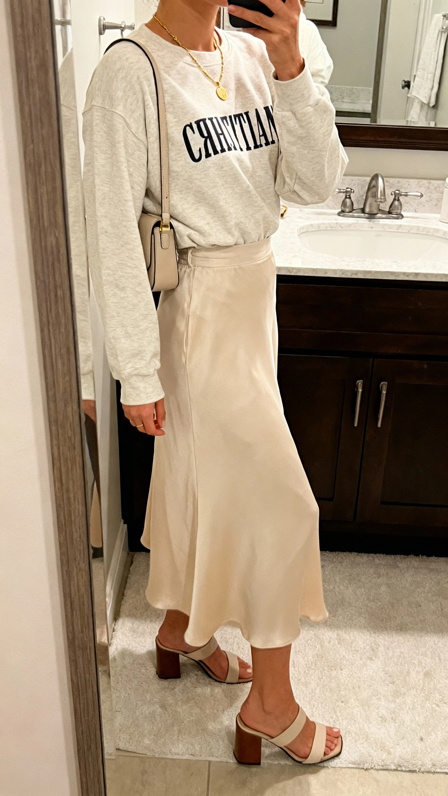 iPhone side-angle mirror selfie of a woman wearing a modest Christian crewneck tucked into a flowy midi skirt, delicate gold necklace, small shoulder bag, and block-heel sandals; face not visible, bathroom mirror, soft indoor lighting, iPhone photo quality.