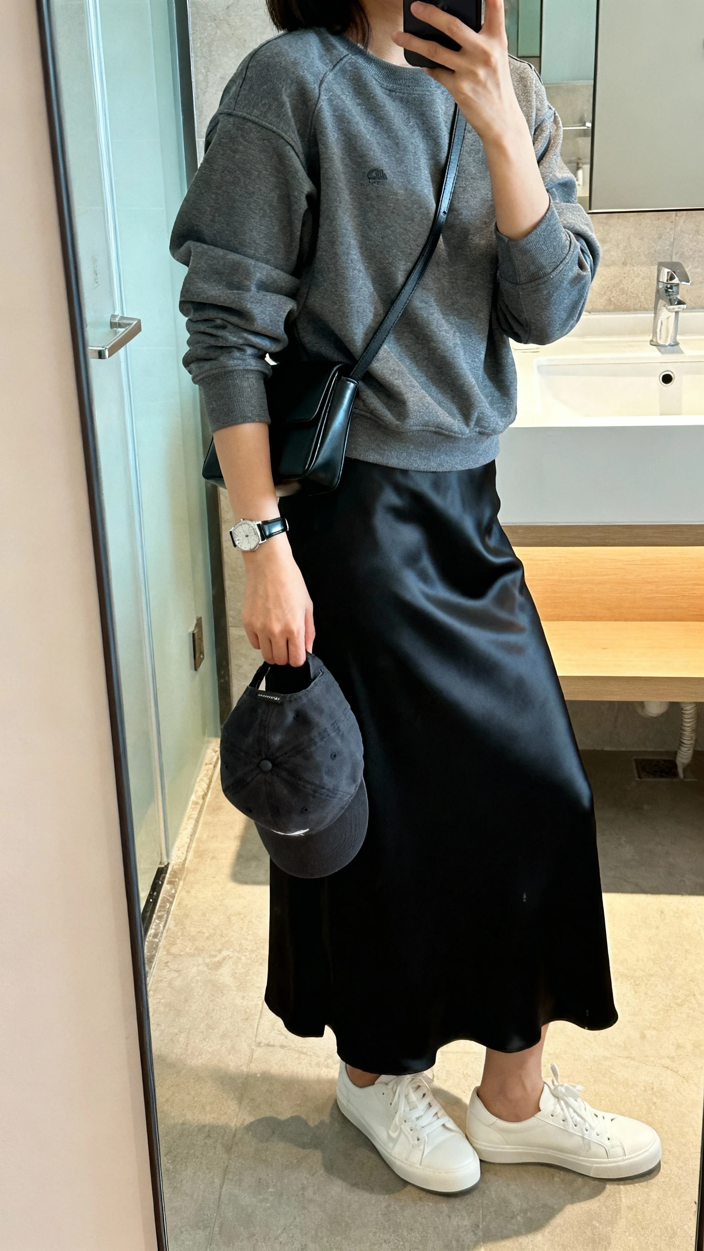 iPhone side-angle mirror selfie of a woman wearing a charcoal sporty crewneck with a satiny black maxi skirt, white low-top sneakers, minimalist black crossbody, sleek watch, and baseball cap in hand, face not visible, bathroom mirror, bright indoor lighting, iPhone photo quality.