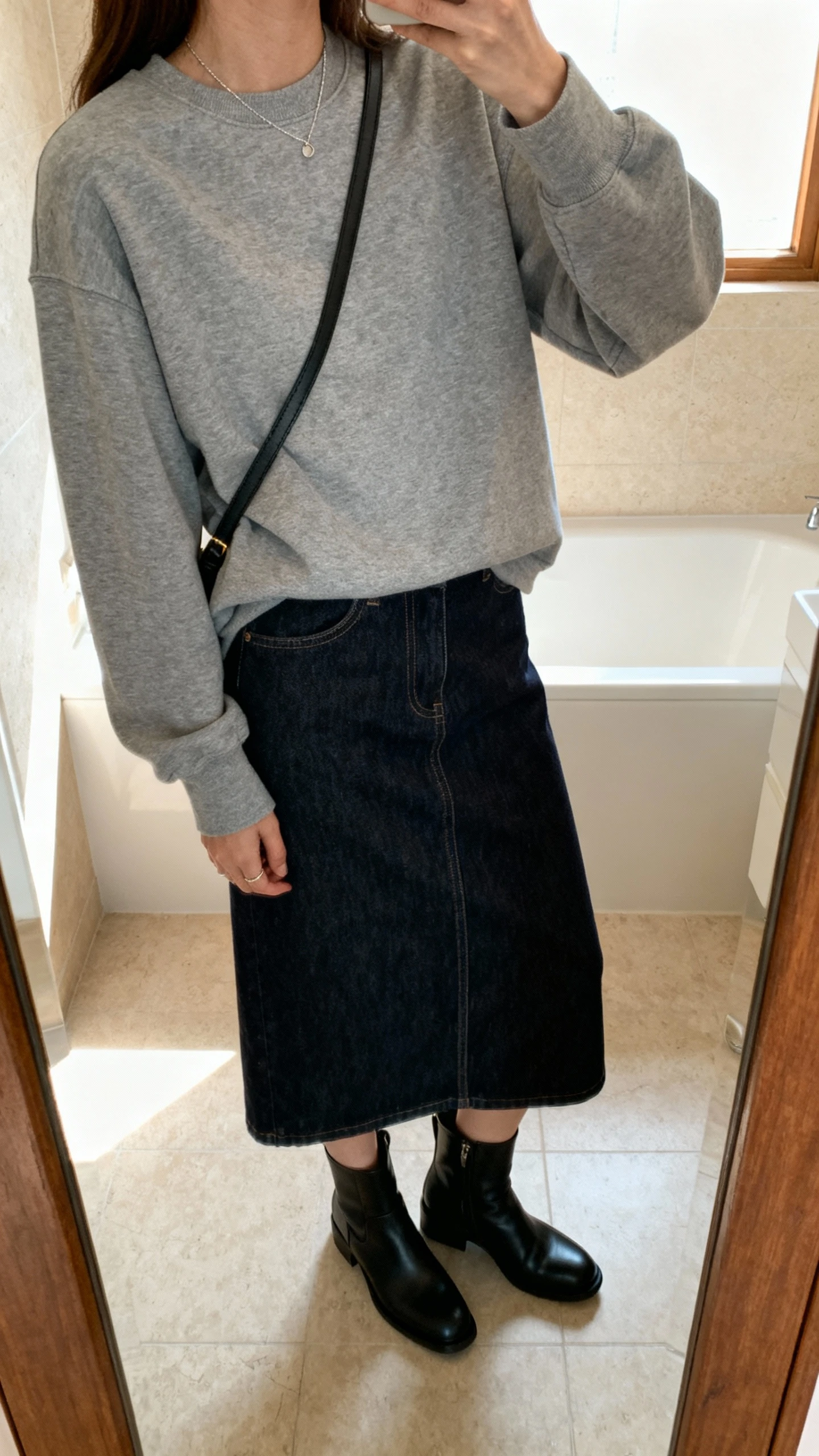 iPhone side-angle mirror selfie of a woman wearing a heather gray crewneck with a dark-wash denim midi skirt, black ankle boots, a simple crossbody bag, and minimal jewelry, face not visible, bathroom mirror, indoor daylight, iPhone photo quality.