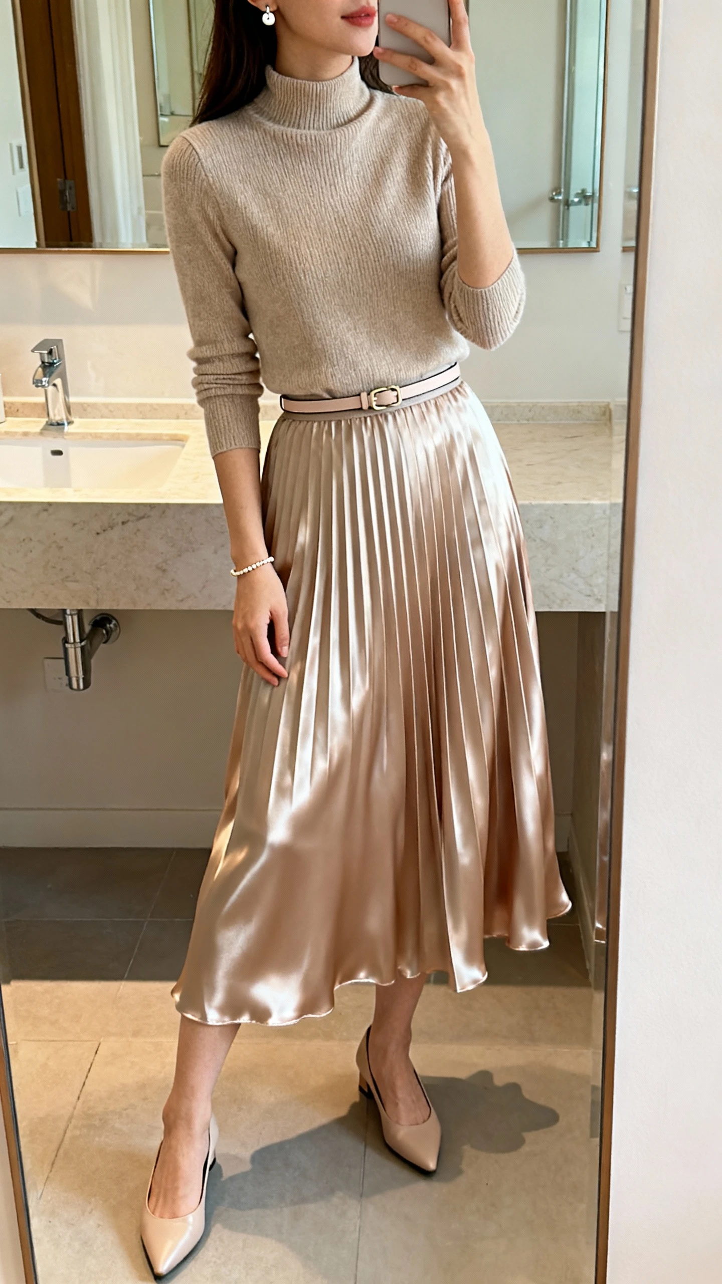 iPhone side-angle mirror selfie of a woman wearing a flowy satin pleated midi skirt, a fitted mock-neck knit top, low kitten heels, a thin waist belt, simple stud earrings, and a dainty bracelet; face not visible, bathroom mirror, soft indoor morning light, iPhone photo quality.
