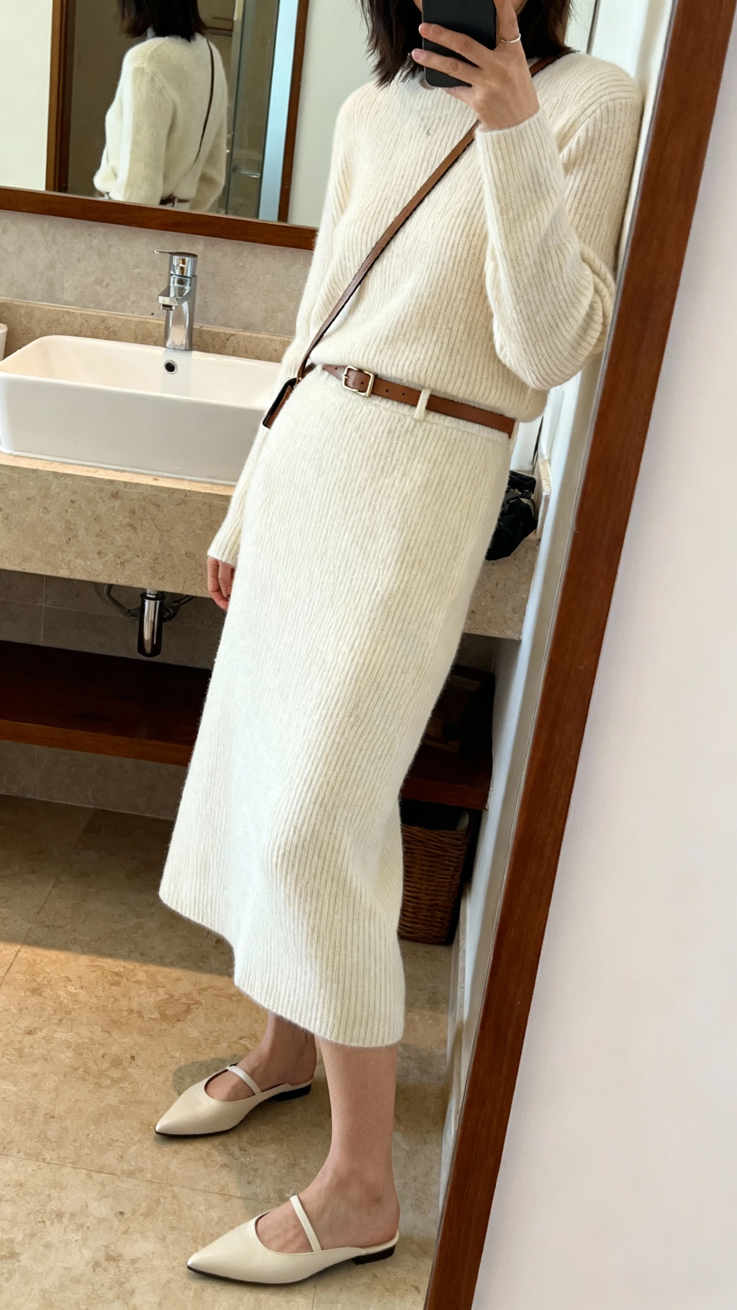 iPhone side-angle mirror selfie of a woman wearing a cream knit midi skirt with a matching fine-gauge sweater, pointed flats, a minimal leather belt, and a slim crossbody bag, face not visible, bathroom mirror, soft indoor lighting, iPhone photo quality.