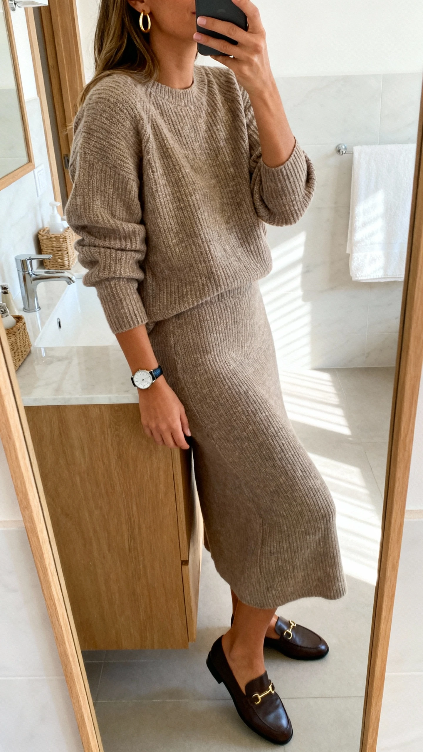 iPhone side-angle mirror selfie of a woman wearing an elevated knit set (matching knit top and midi skirt) with minimal gold hoops, sleek watch, and loafers, face not visible, bathroom mirror, soft morning light, iPhone photo quality.