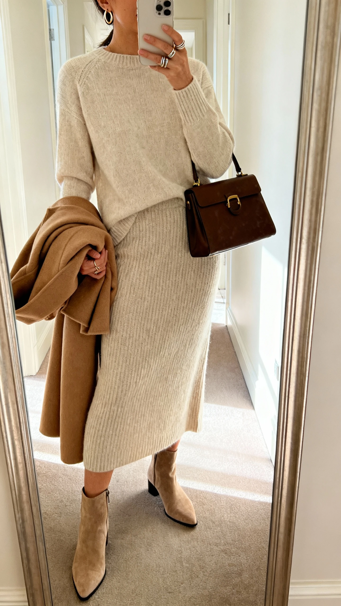 iPhone side-angle mirror selfie of a woman wearing a fine-gauge crewneck sweater with a coordinating knit midi skirt, almond-toe ankle boots, subtle hoop earrings, stackable rings, and a structured satchel, with an optional camel wool coat folded over her arm, face not visible, cropped at shoulders, hallway mirror, morning light, iPhone photo quality.