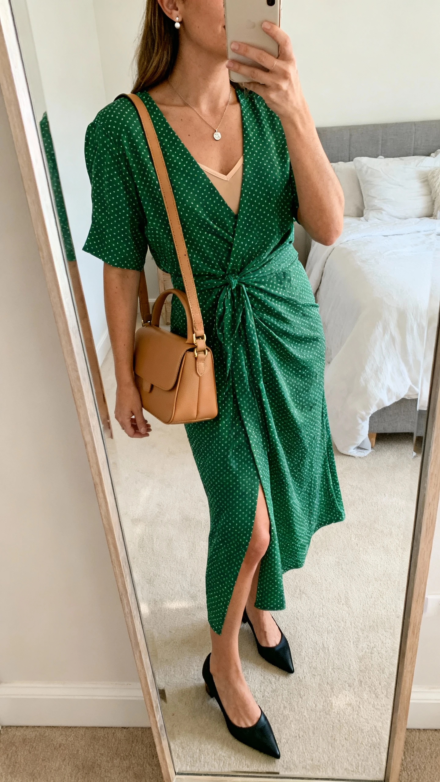 iPhone side-angle mirror selfie of a woman wearing a forest green wrap polka dot dress, pointed-toe kitten heels in black, a structured camel shoulder bag, a dainty pendant necklace, stud earrings, and a lightweight skin-tone slip underneath, face not visible, cropped at shoulders, bedroom mirror, soft morning light, iPhone photo quality.