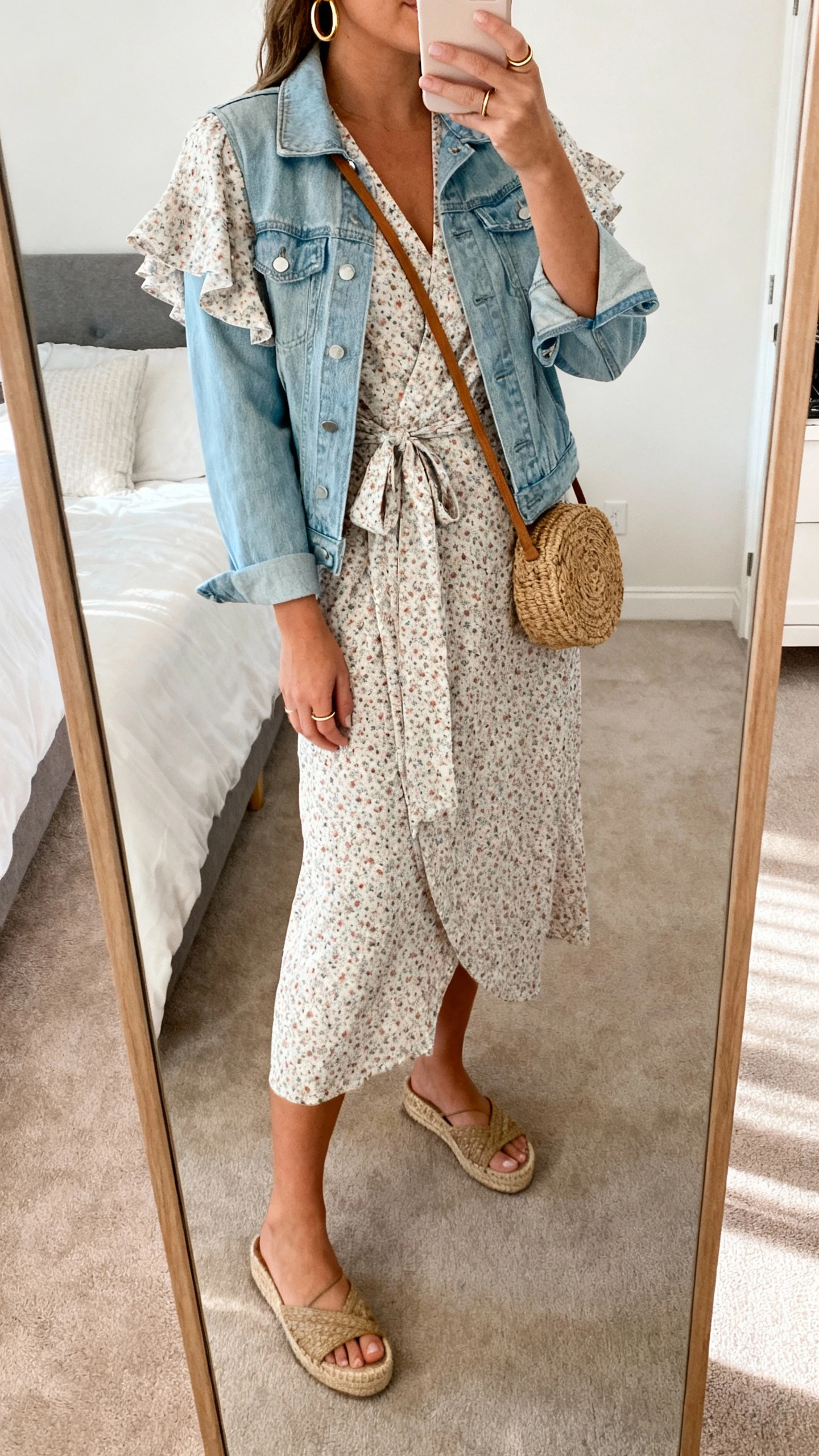 iPhone side-angle mirror selfie of a woman wearing a wrap midi dress with tiny blooms, V-neck and flutter sleeves, a soft denim jacket, espadrille wedges, a woven crossbody bag, gold hoops, a slim stacking ring, and a slip peeking subtly; face not visible, bedroom full-length mirror, natural daylight, iPhone photo quality.