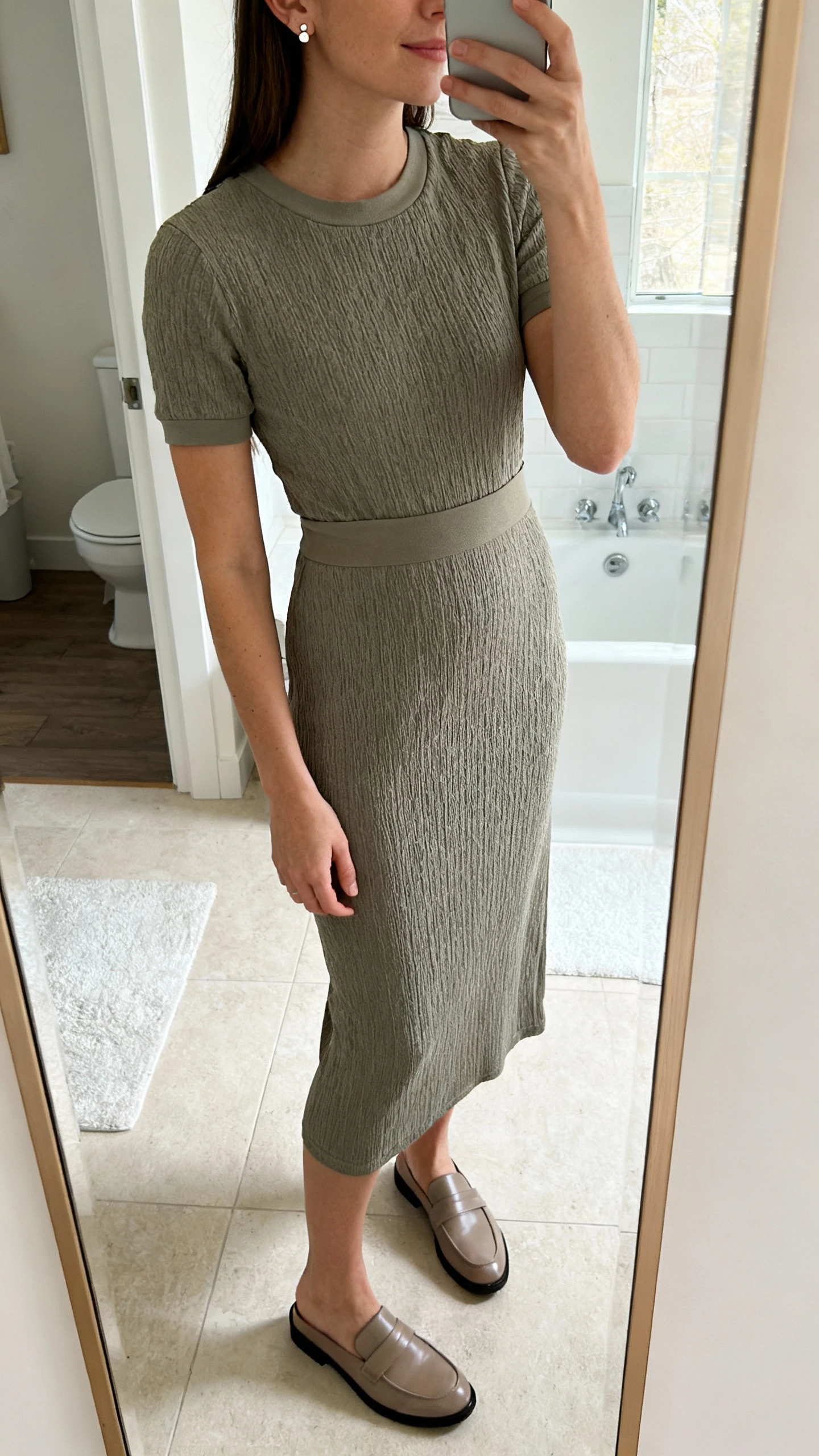 iPhone side-angle mirror selfie of a woman wearing a stretch knit textured dress in a midi length with a clean crew neckline and fitted waist, paired with simple stud earrings and loafers; face not visible, cropped at shoulders, bathroom mirror, bright indoor lighting, iPhone photo quality.