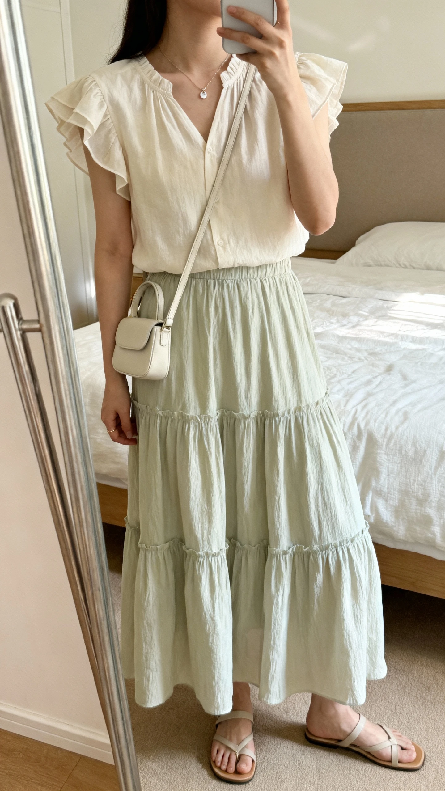 iPhone side-angle mirror selfie of a woman wearing a flutter-sleeve blouse with a tiered maxi skirt, lightweight crossbody bag, dainty pendant, flat sandals, face not visible, cropped at shoulders, bedroom mirror, soft afternoon light, iPhone photo quality.