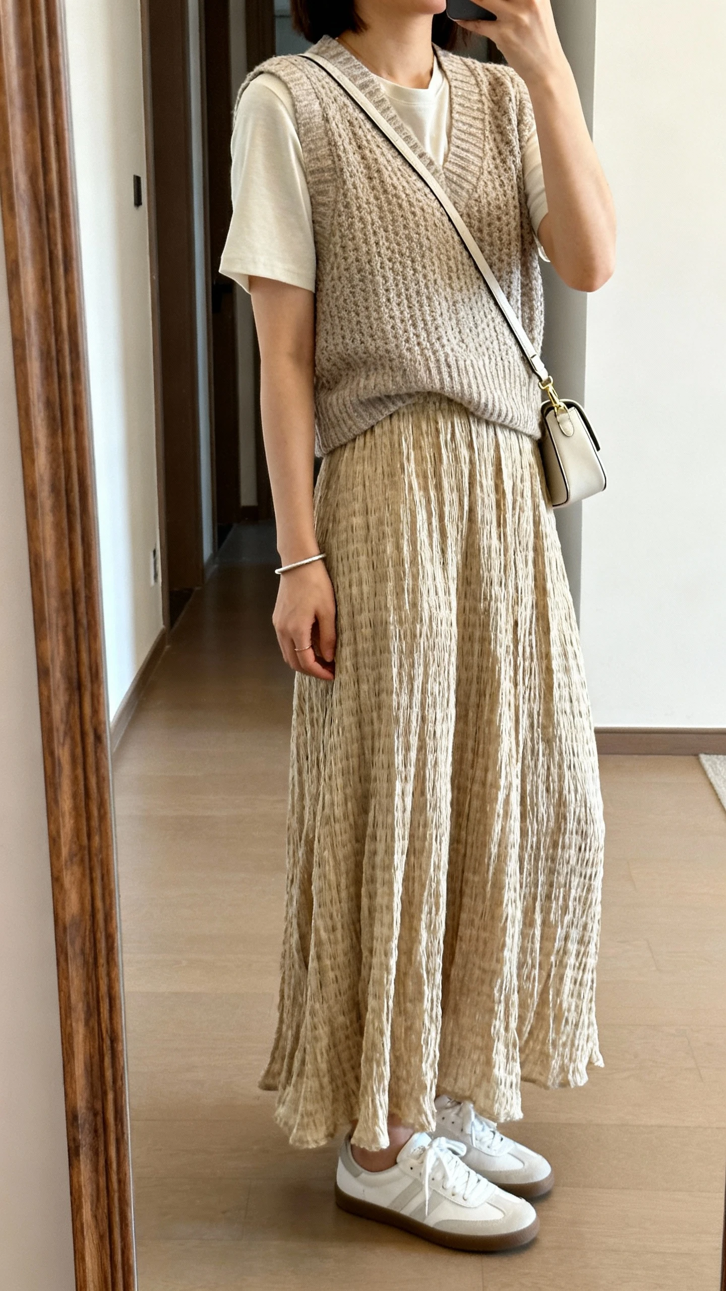 iPhone side-angle mirror selfie of a woman wearing a knit vest over a lightweight tee with a flowing woven maxi skirt, white sneakers, slim bracelet, and a mini shoulder bag; face not visible, cropped at shoulders, hallway mirror with soft natural light, iPhone photo quality.