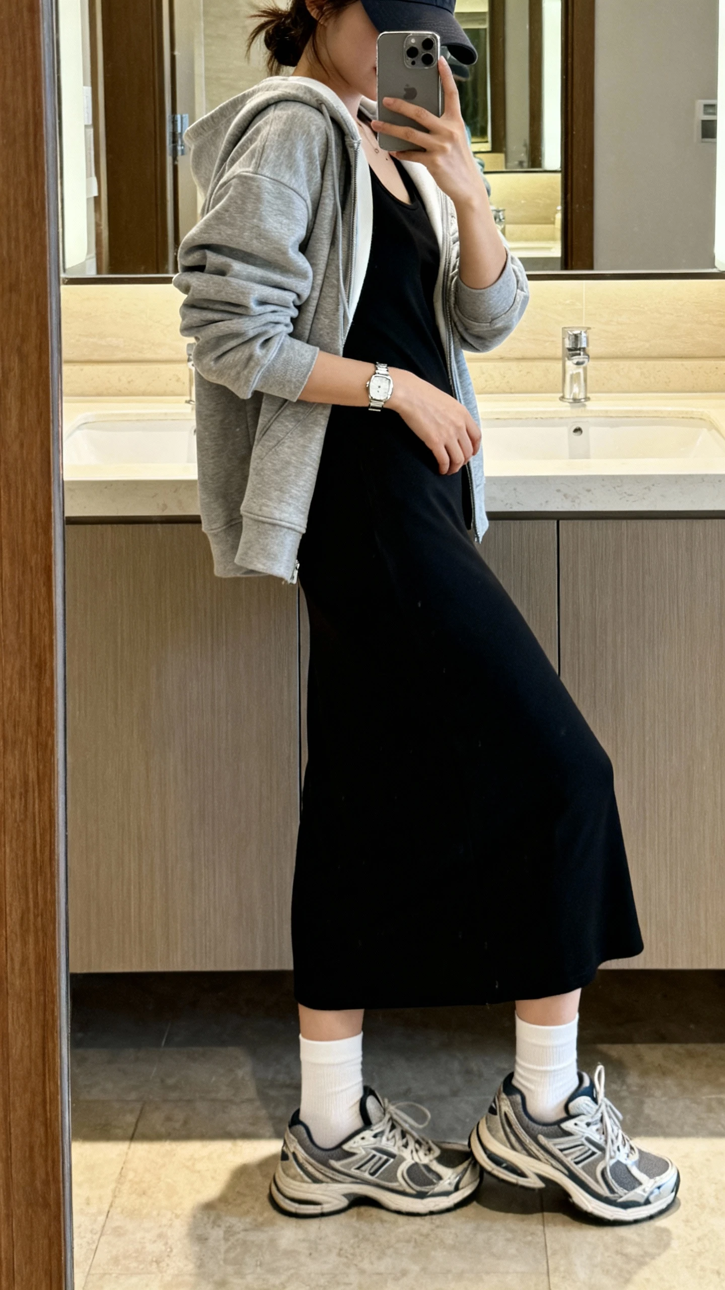 iPhone side-angle mirror selfie of a woman wearing a sporty-casual hoodie layered over a sleek black maxi dress, white crew socks, retro running sneakers, baseball cap, and a minimal silver watch, face not visible, cropped at shoulders, bathroom mirror, soft indoor lighting, iPhone photo quality.