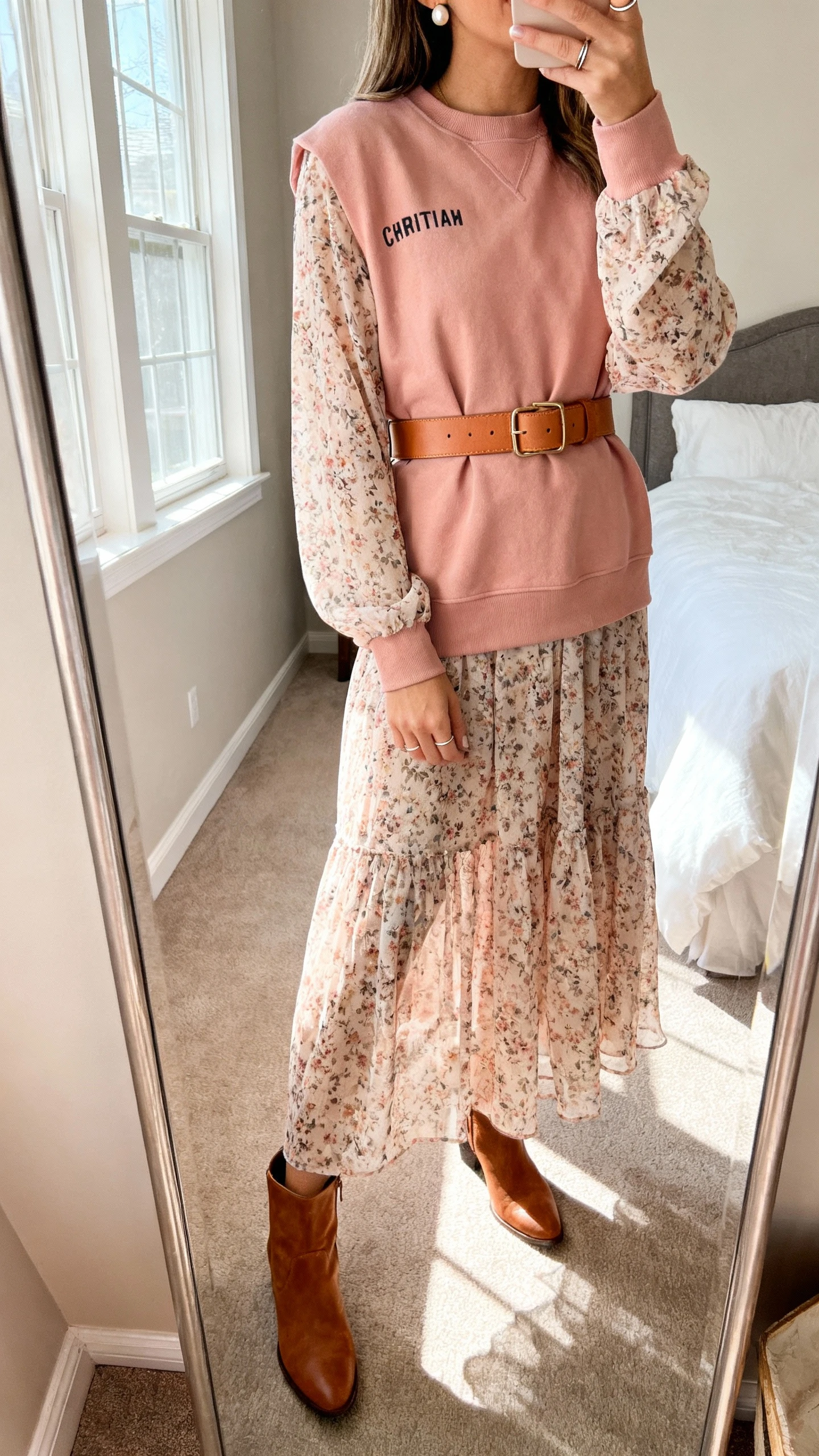 iPhone side-angle mirror selfie of a woman wearing a blush Christian crewneck belted with a tan leather waist belt over a long-sleeve floral chiffon maxi dress, cognac ankle boots, pearl studs, and a thin stacking ring, face not visible, cropped at shoulders, bedroom mirror, afternoon window light, iPhone photo quality.