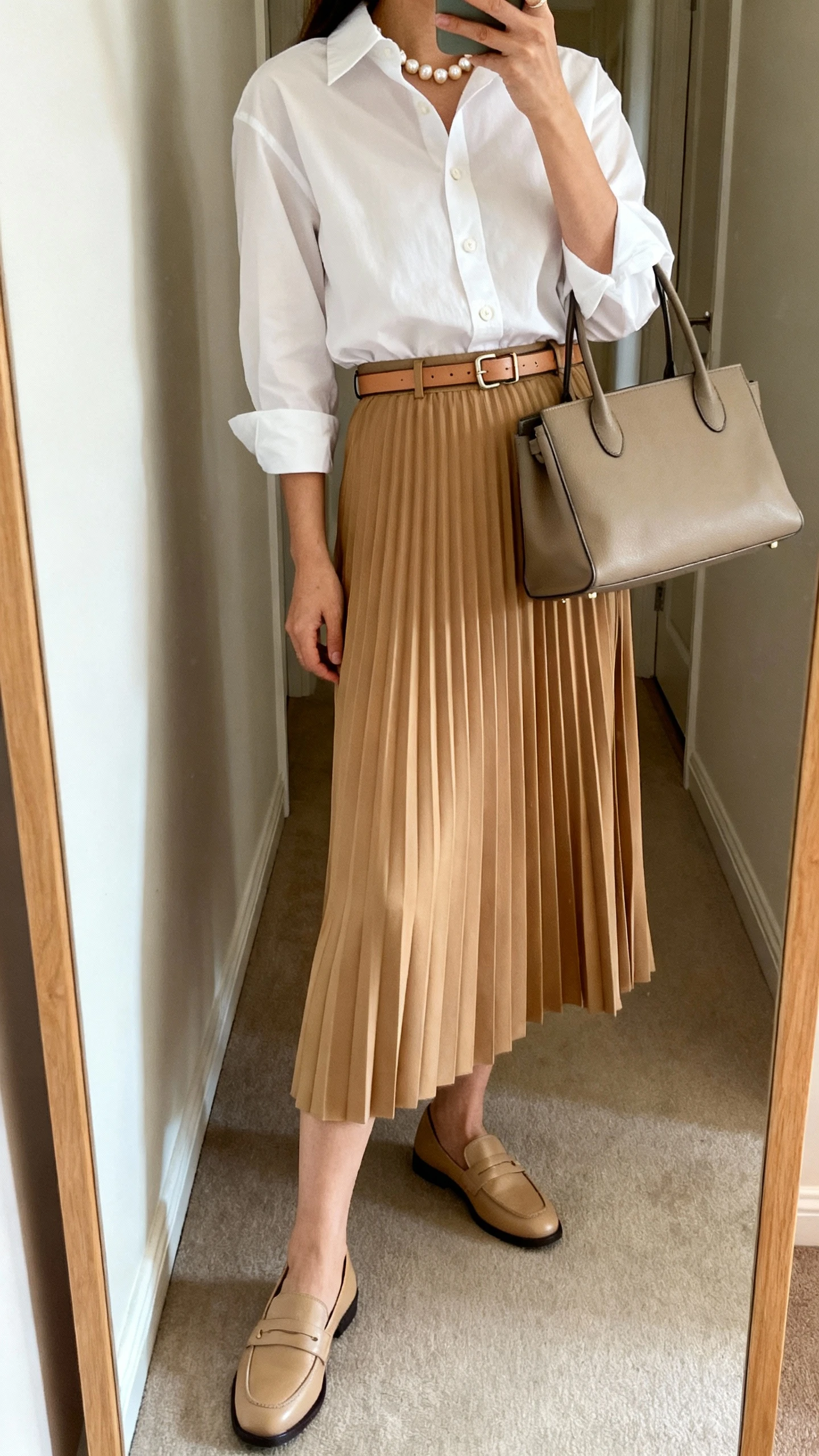 iPhone side-angle mirror selfie of a woman wearing a pleated midi skirt in camel with a crisp white button-up tucked in, thin leather belt, classic loafers, pearl studs, and a structured tote, face not visible, cropped at shoulders, full-length mirror in hallway, soft natural daylight, iPhone photo quality.