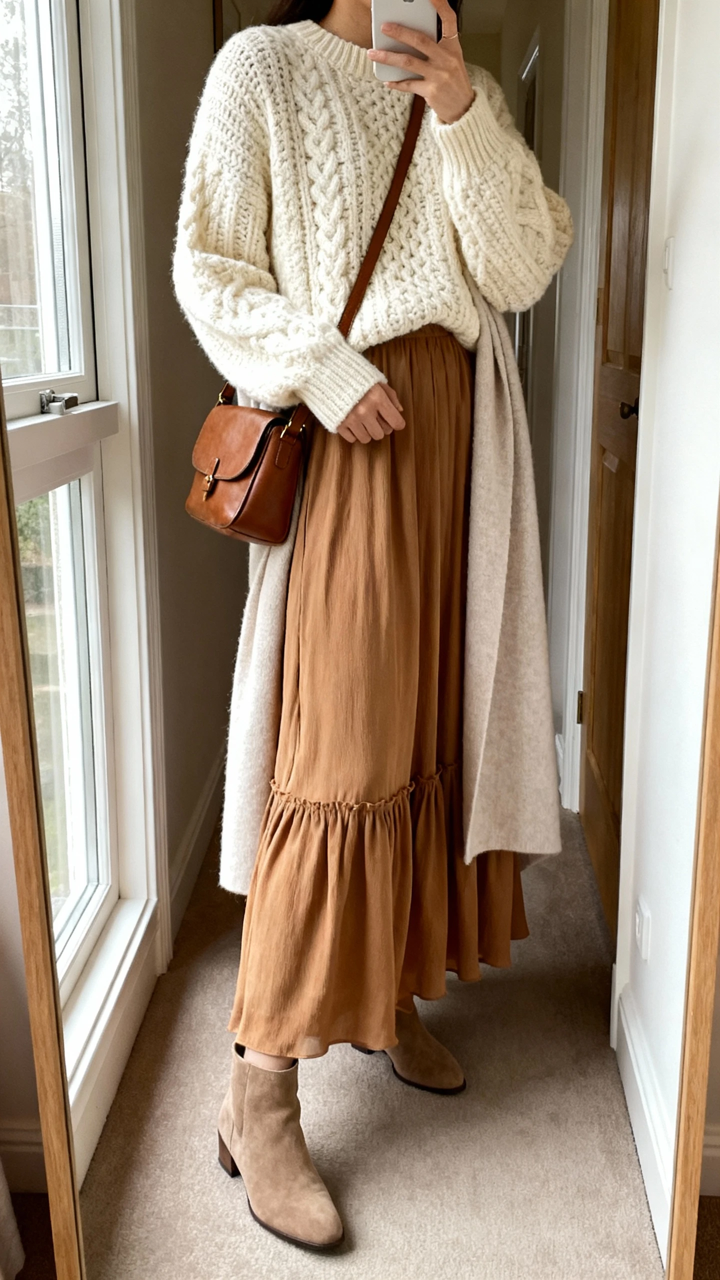 iPhone side-angle mirror selfie of a woman wearing a flowy lined maxi skirt with a chunky knit sweater in cream or camel, minimal ankle boots, a crossbody leather bag, and a light longline cardigan or wrap, face not visible, cropped at shoulders, full-length mirror in hallway, soft window light, iPhone photo quality.