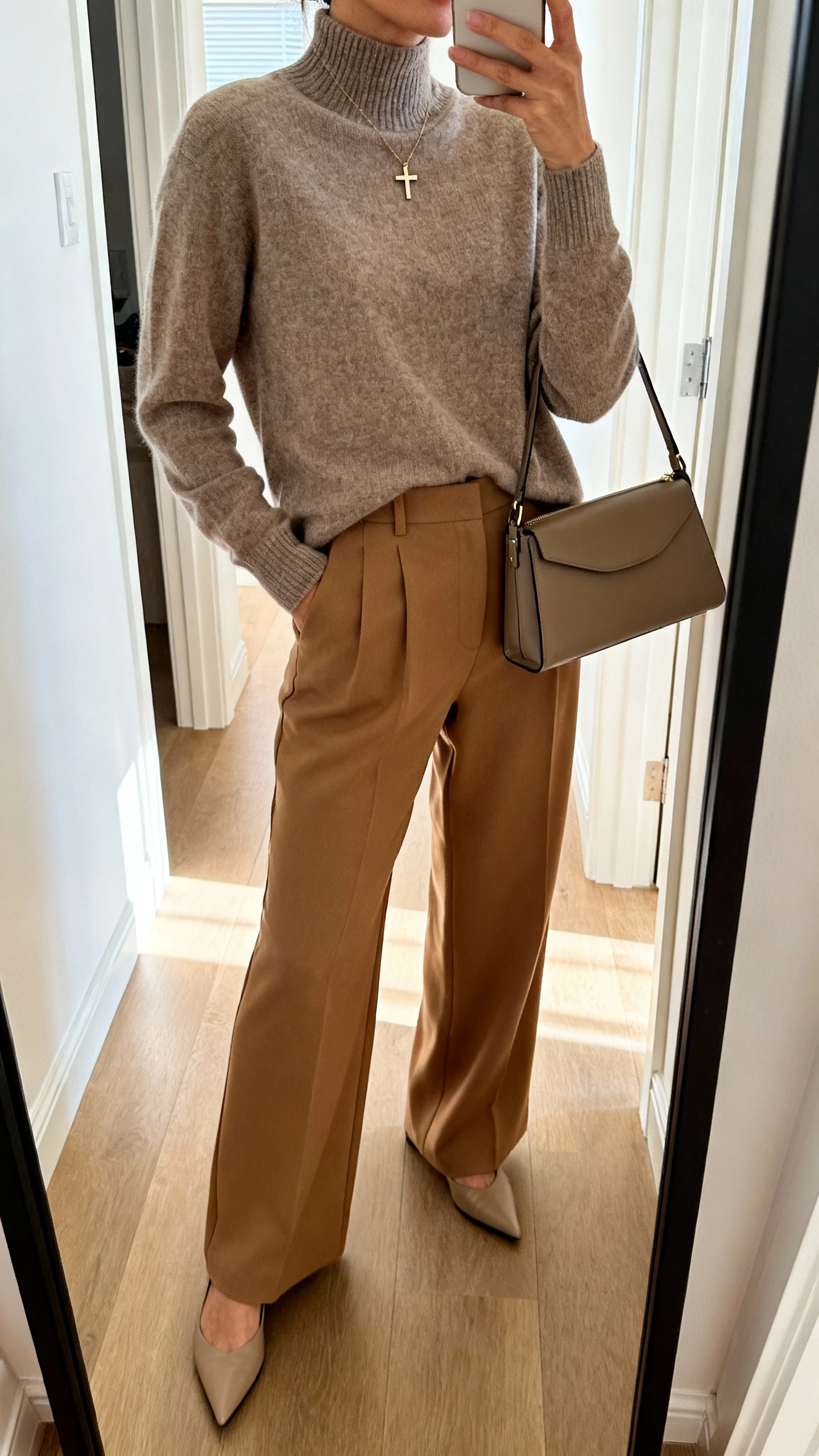 iPhone side-angle mirror selfie of a woman wearing a taupe lightweight mock-neck sweater with high-waisted camel wide-leg trousers, pointed flats, a small cross pendant necklace, and a structured satchel, face not visible, cropped at shoulders, full-length mirror in hallway, soft indoor daylight, iPhone photo quality.