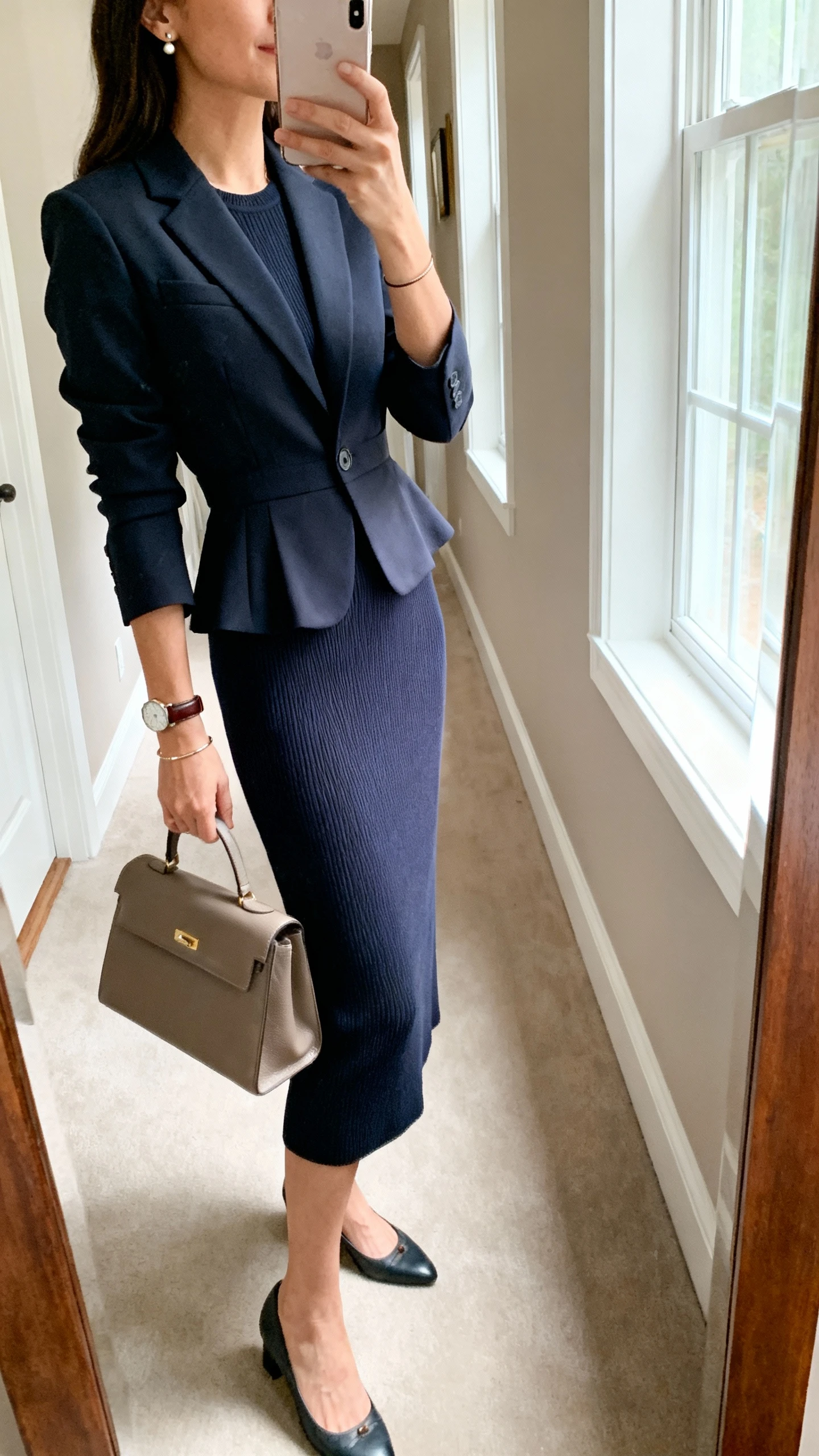 iPhone side-angle mirror selfie of a woman wearing a midi sheath dress with a modest neckline in sturdy ponte/structured knit, a tailored single-breasted blazer nipped at the waist, classic pumps or low slingbacks, a leather watch, thin bracelet, stud earrings, and a medium structured handbag; face not visible, cropped at shoulders, hallway mirror with natural window light, iPhone photo quality.
