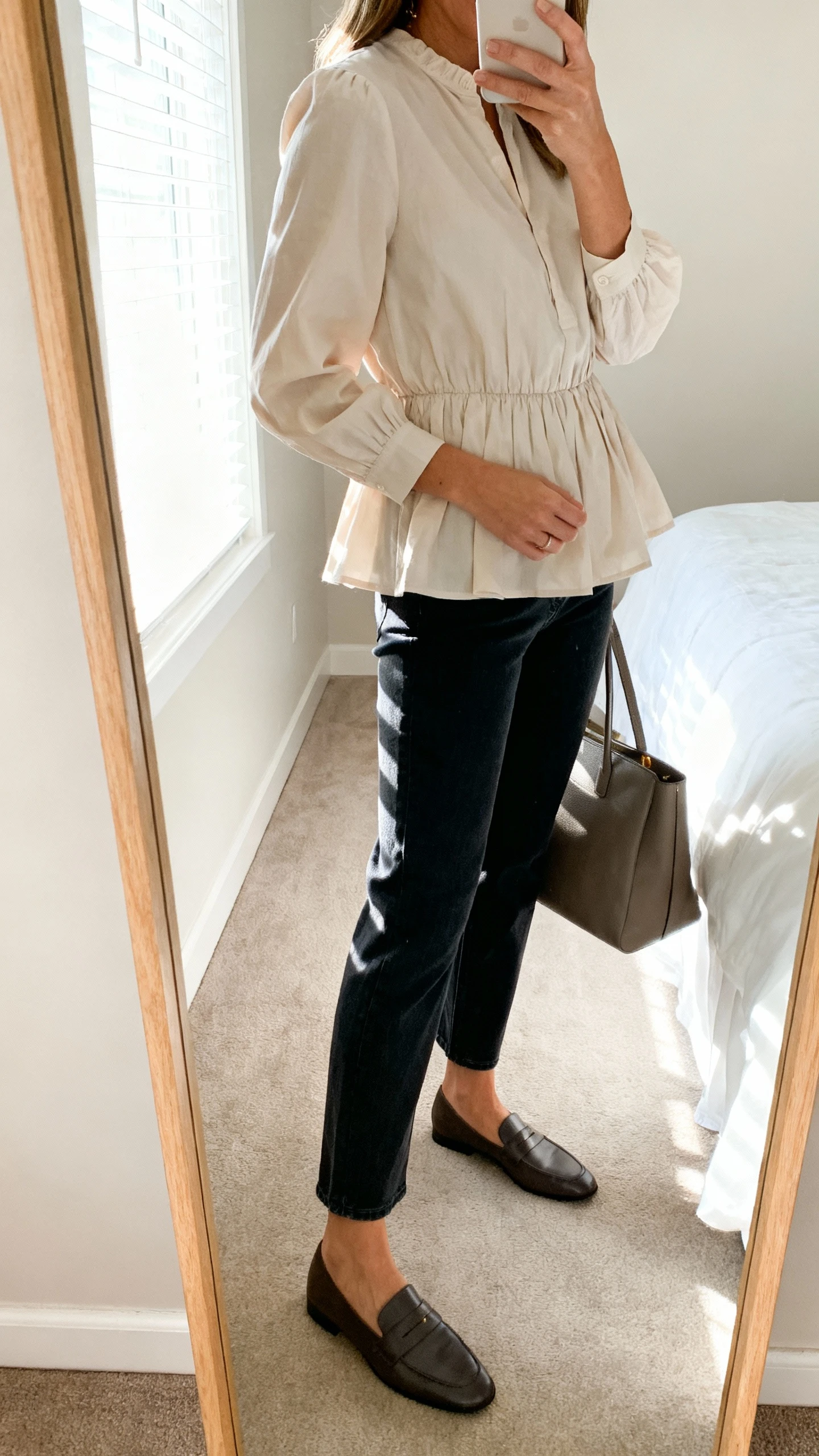 iPhone side-angle mirror selfie of a woman wearing a soft poplin peplum blouse that gently defines the waist, with straight-leg dark jeans and loafers, carrying a structured tote; face not visible, bedroom mirror, bright window light, iPhone photo quality.