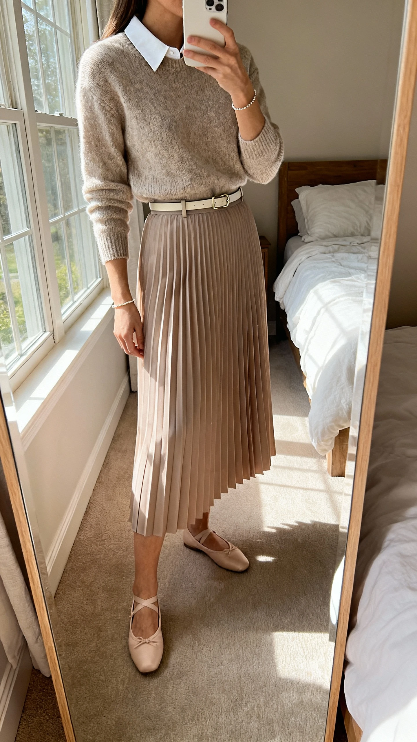 iPhone side-angle mirror selfie of a woman wearing a pleated skirt with a collared knit top tucked in, ballet flats, a thin belt, and a delicate bracelet, face not visible, bedroom mirror, afternoon window light, iPhone photo quality.